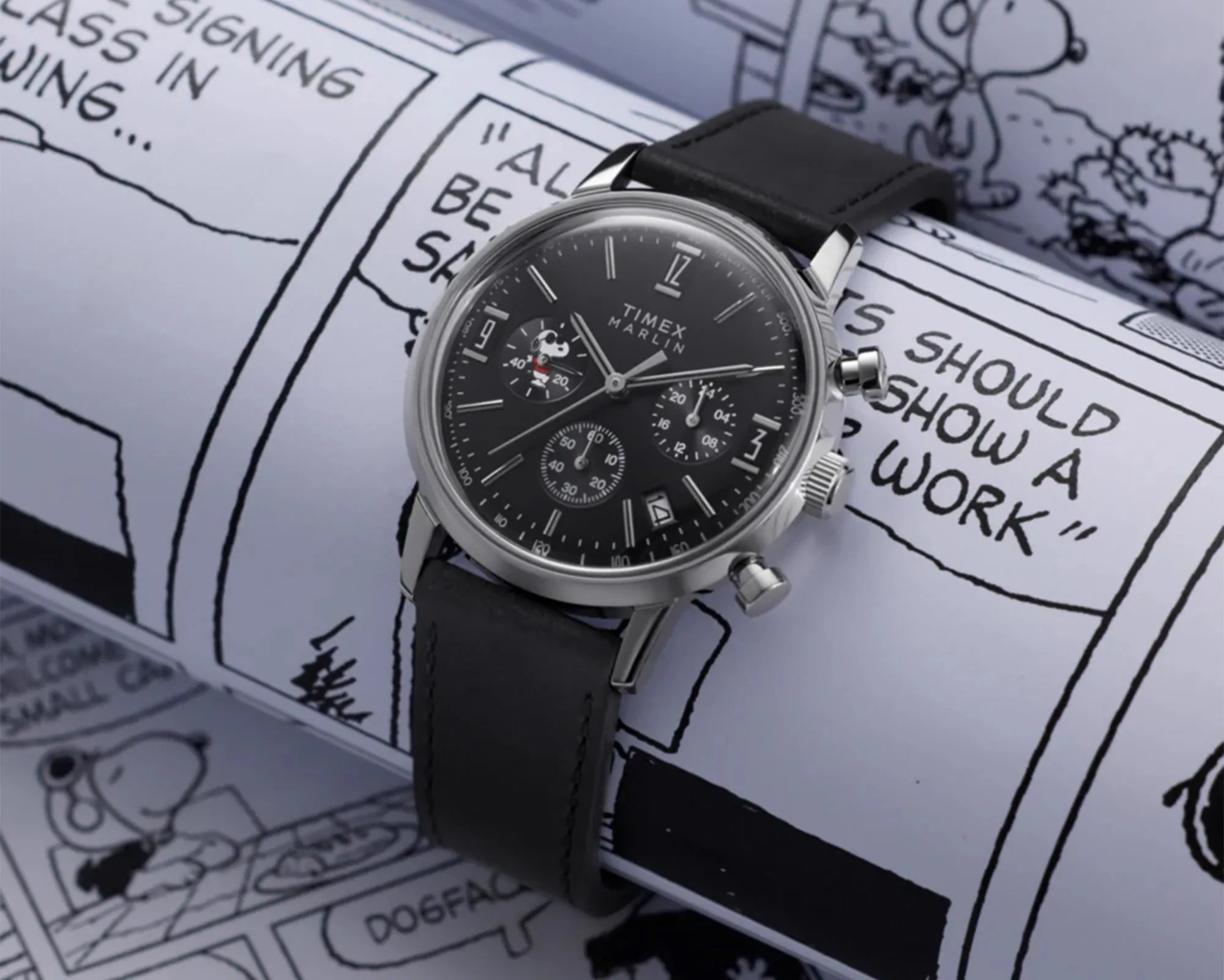 Timex Marlin black chronograph watch with Snoopy illustration on dial and black leather strap on comic strip background.