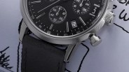 Close-up of a black chronograph watch face with silver markers, date window showing 14, and black leather strap.