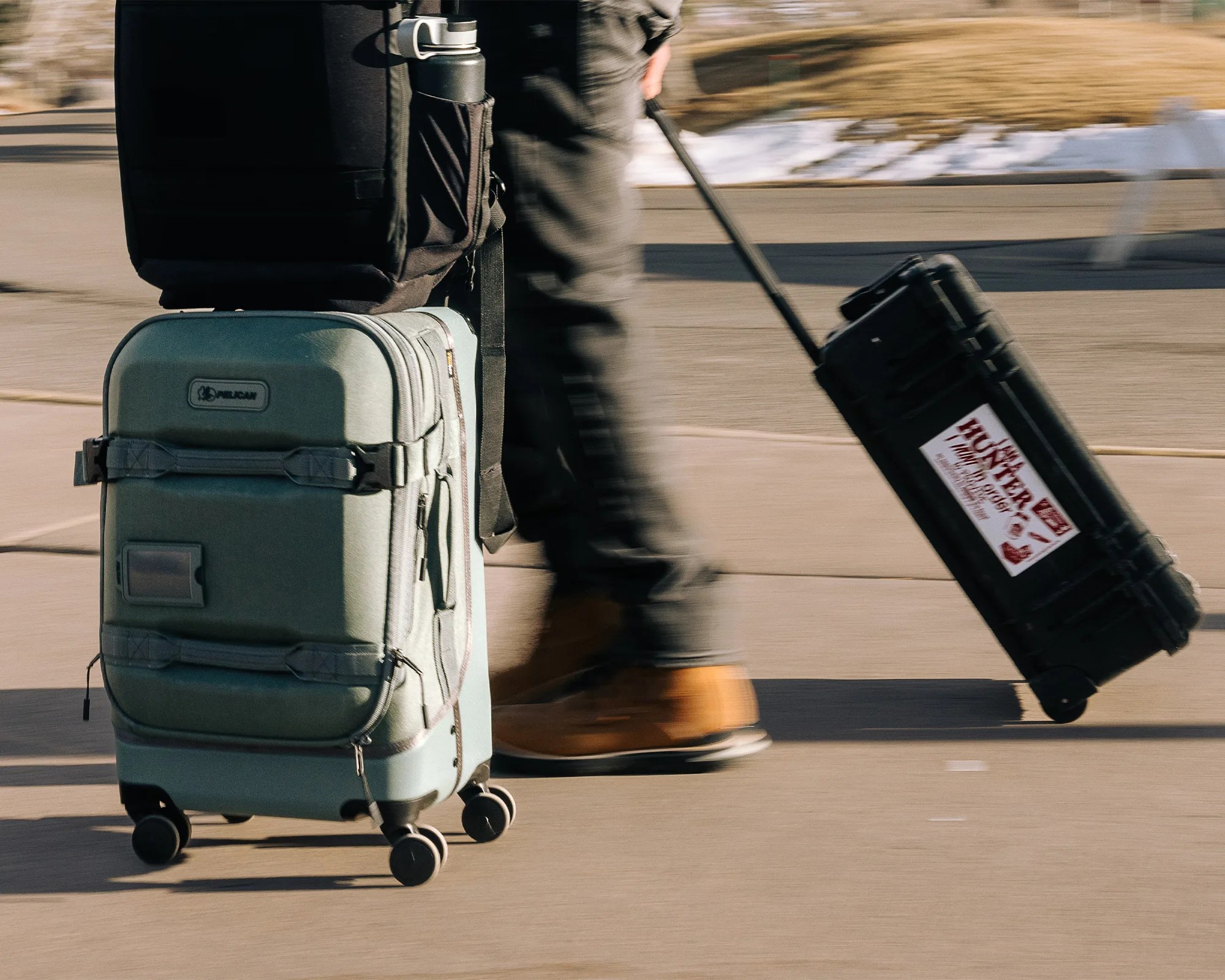 Person walking on pavement pulling a black hard case with a sticker and rolling a green soft suitcase with four wheels.