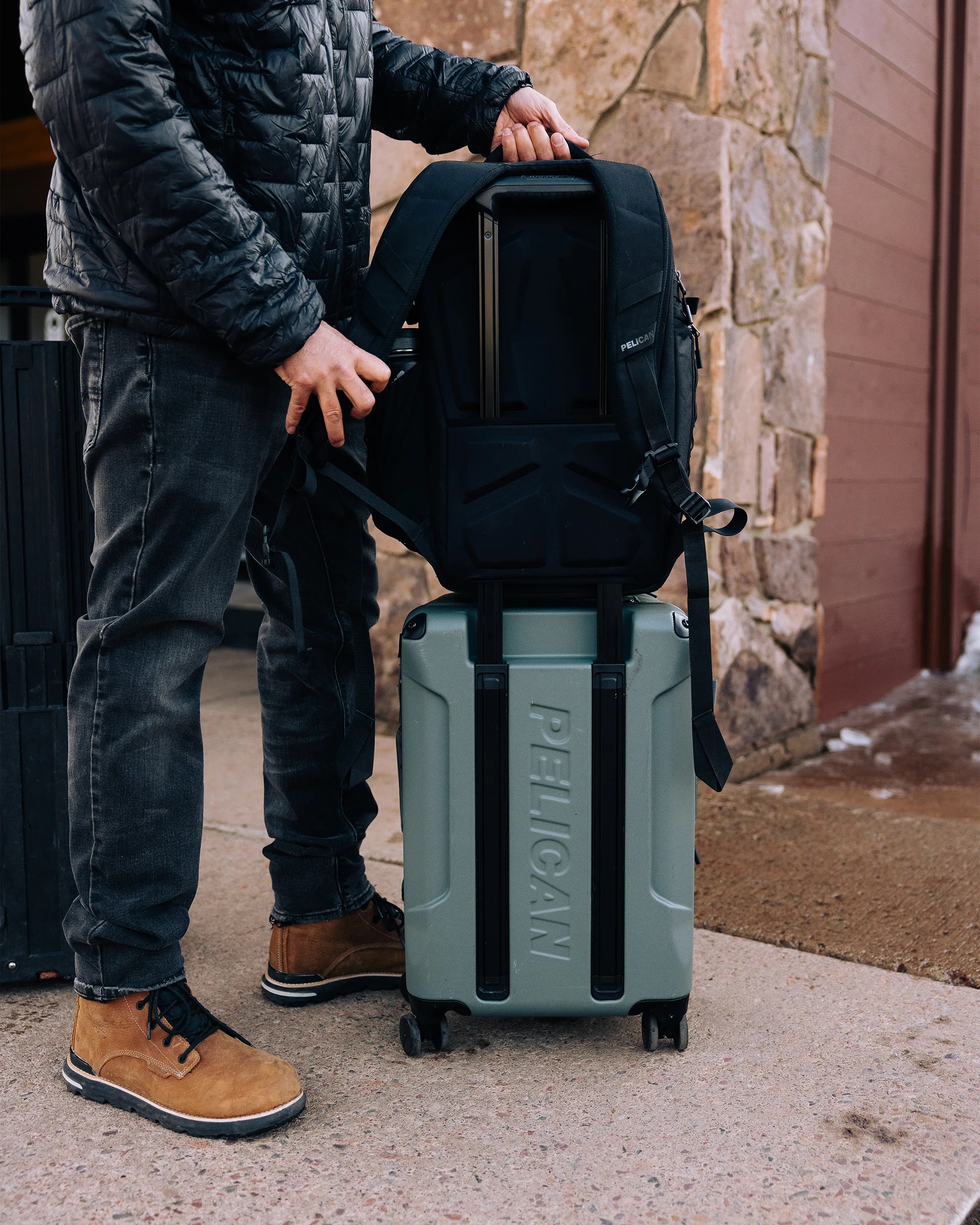 Person wearing brown boots and black jeans holding a black Pelican backpack on top of a green Pelican rolling suitcase.