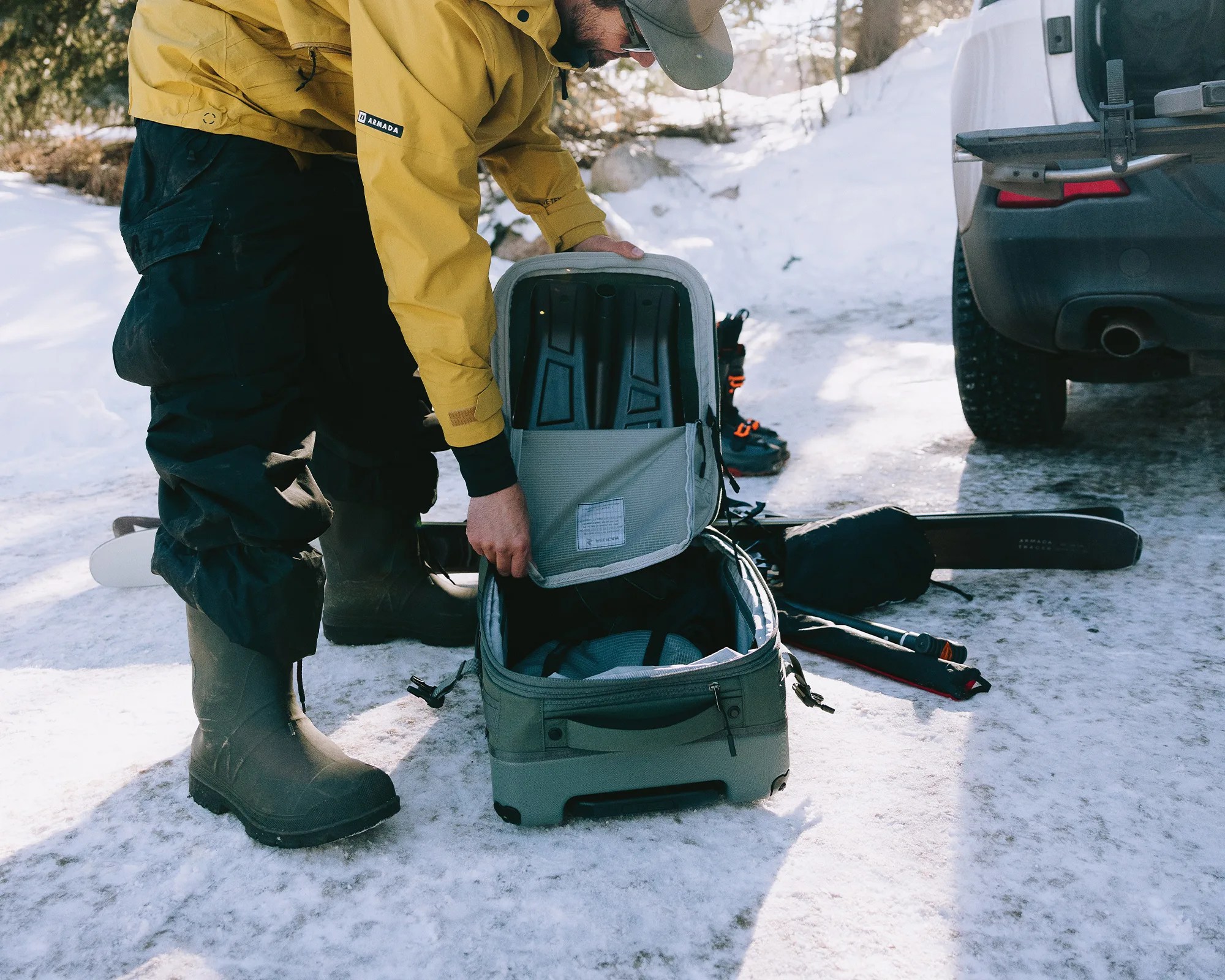 Person in yellow jacket and black snow pants packing black ski boots into a gray suitcase on snow next to skis and a vehicle.