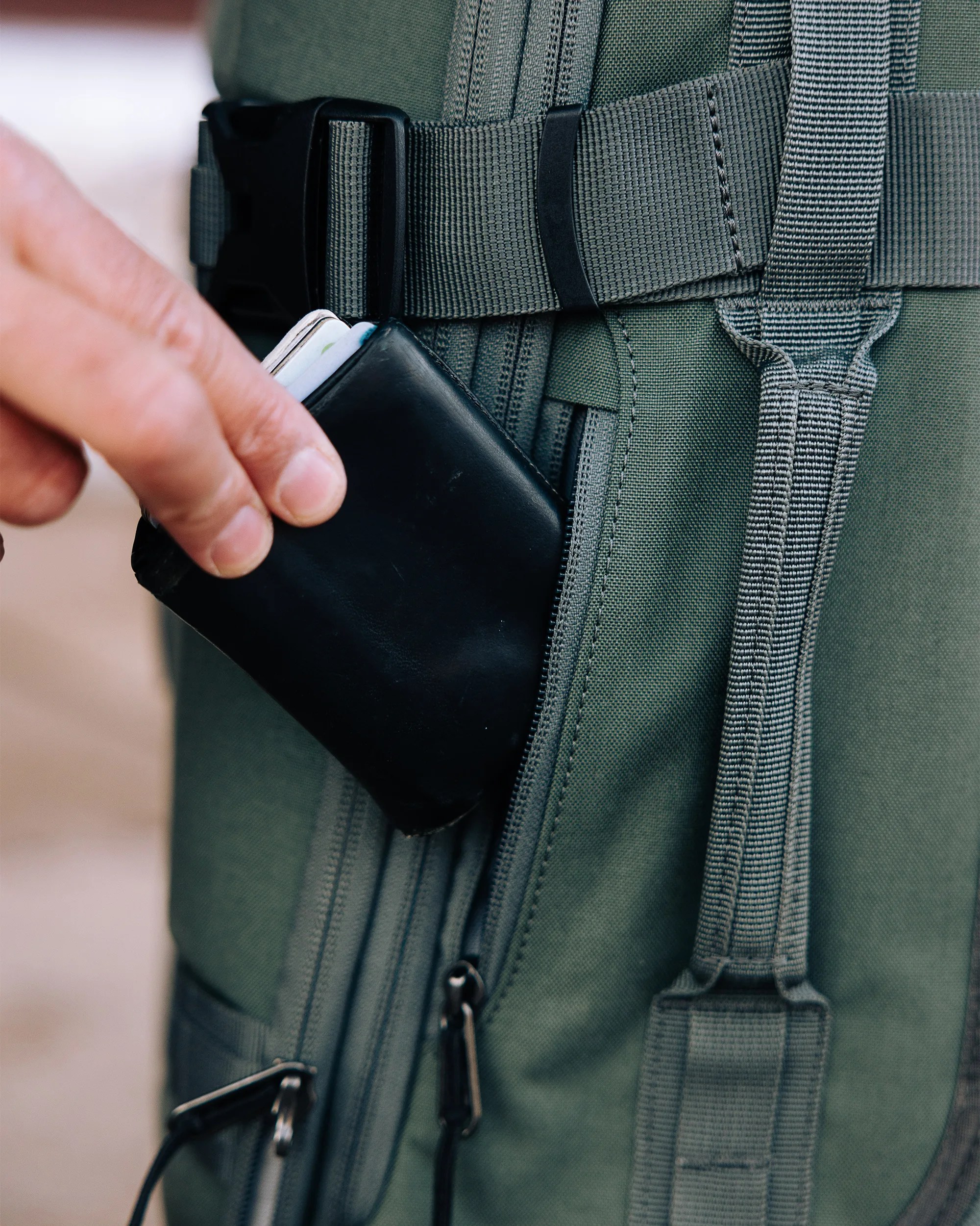 Hand placing a black leather wallet into the zippered pocket of an olive green backpack.