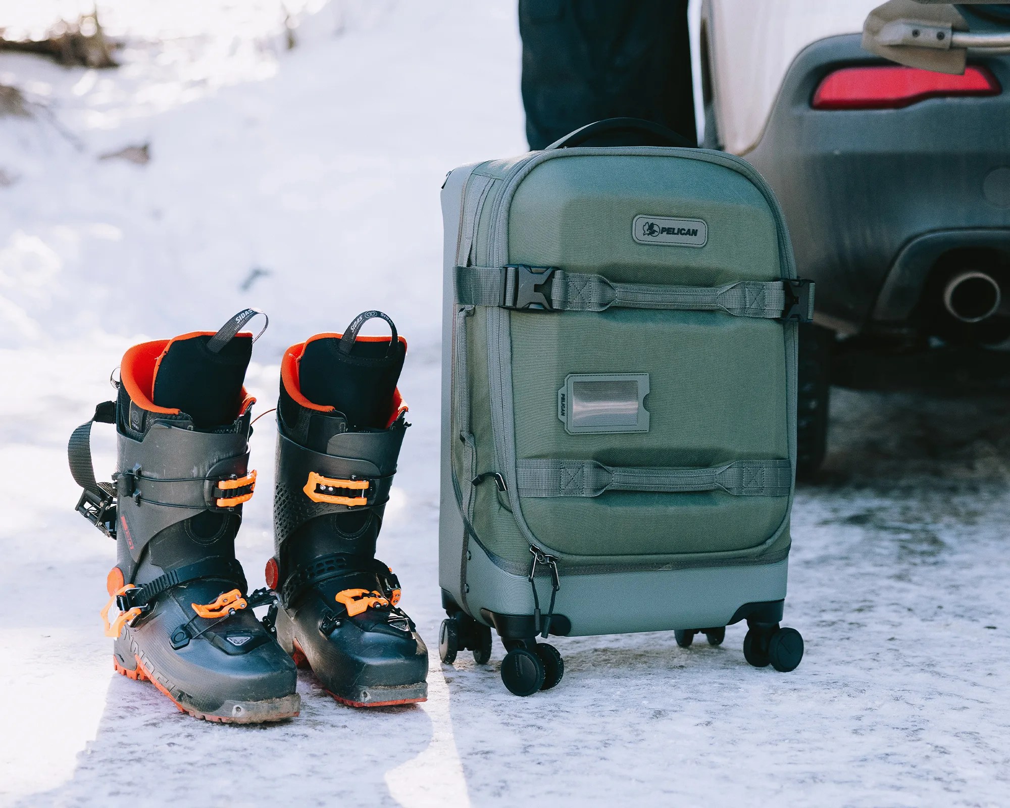 Pair of black and orange ski boots next to a green Pelican rolling suitcase on snowy ground.