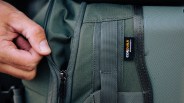 Hand holding the zipper of a green Cordura fabric bag with black webbing straps.