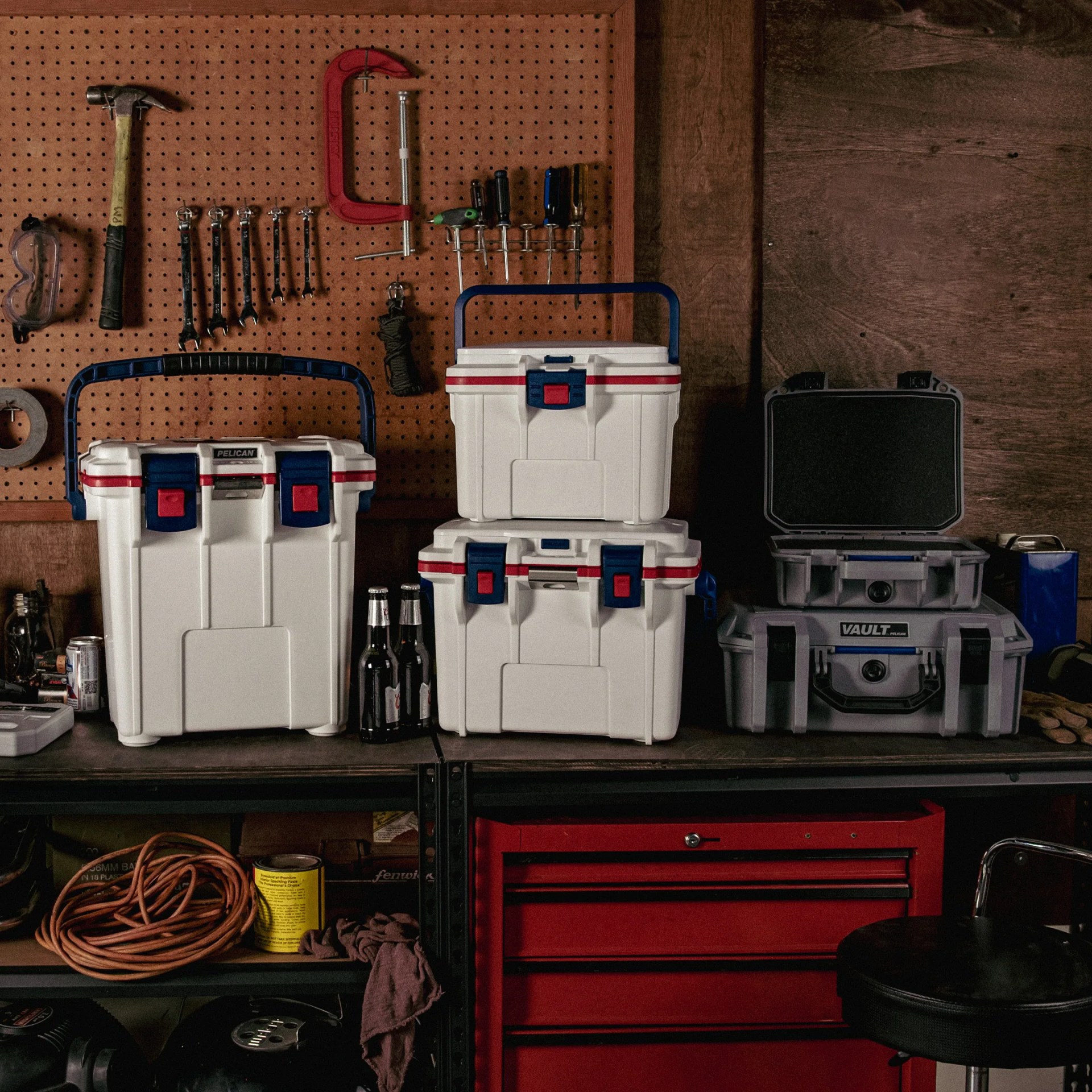 Three white Pelican coolers with blue handles and red latches on a workbench with tools and a red toolbox.