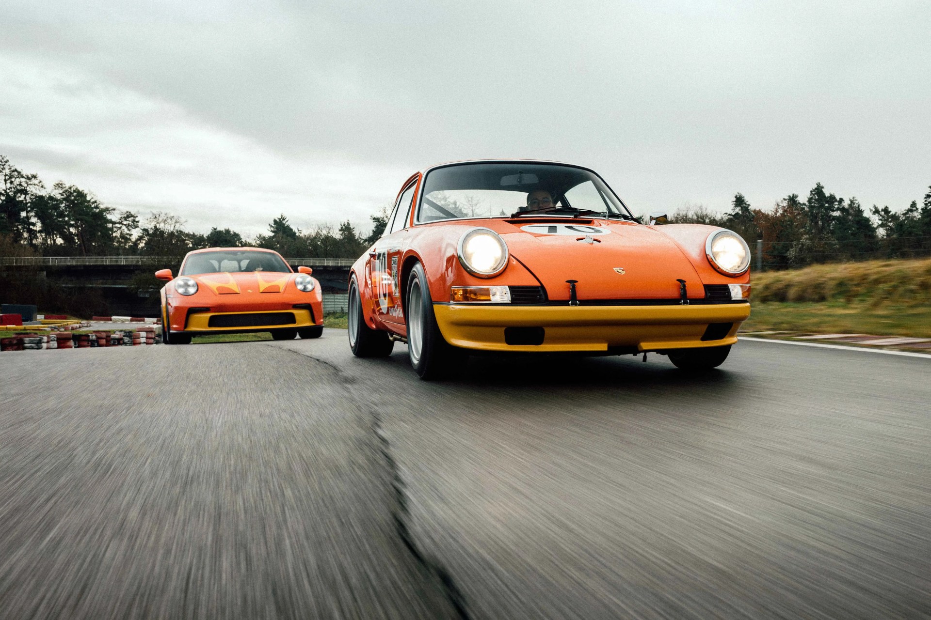 Two orange Porsche race cars with yellow accents driving on a racetrack under an overcast sky.