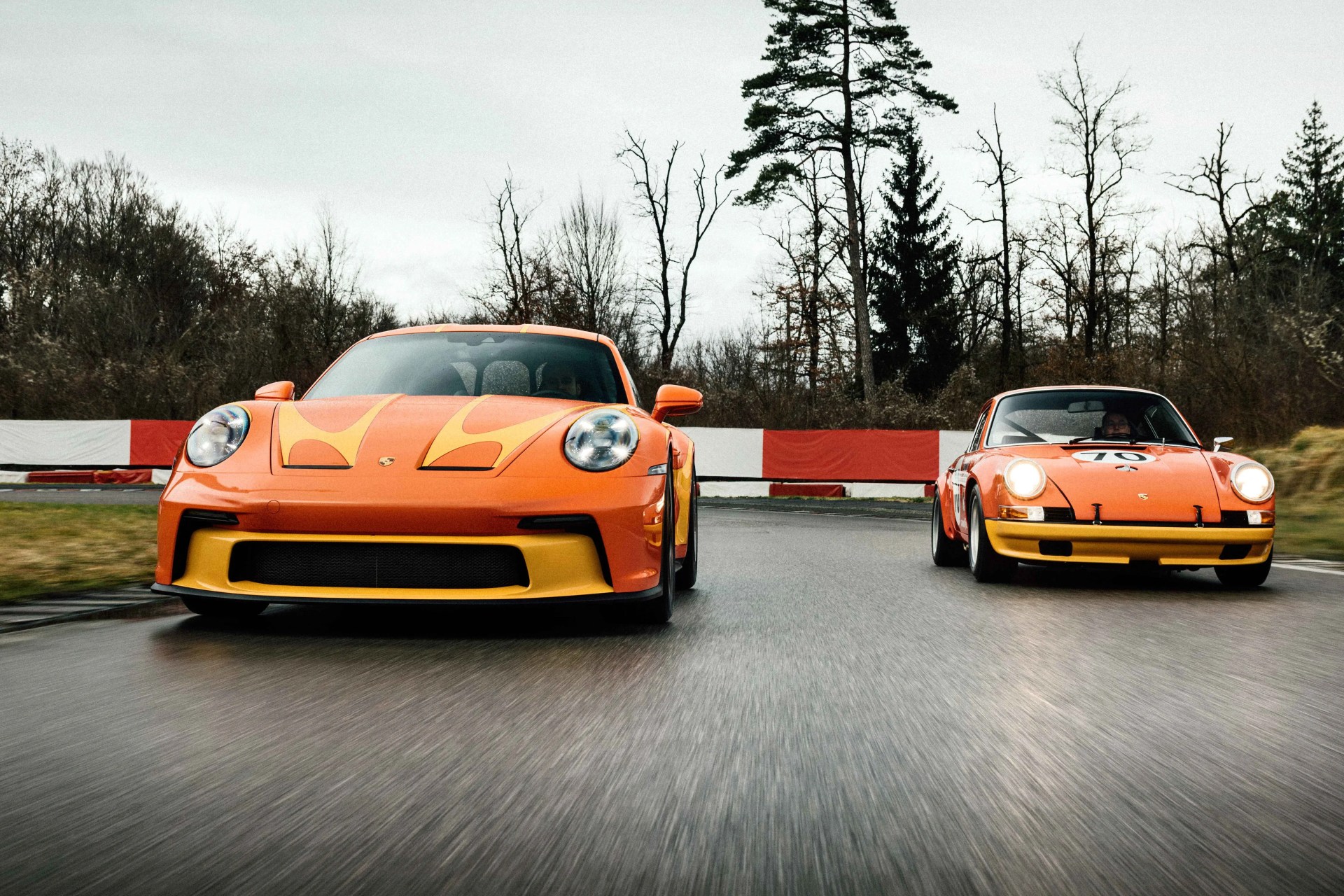 Two orange Porsche sports cars with yellow accents driving on a wet racetrack with bare trees in the background.