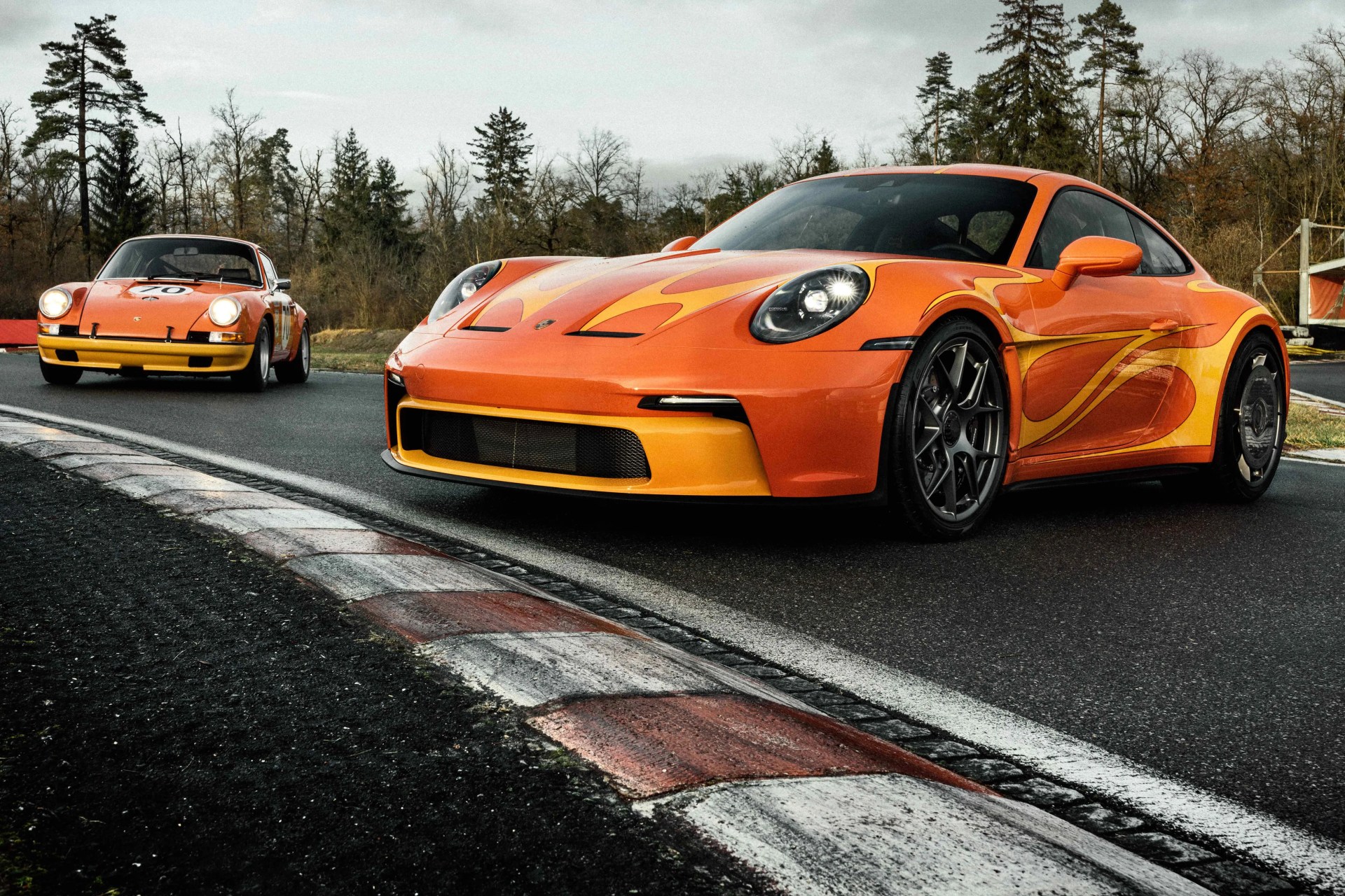 Two orange Porsche sports cars with yellow flame decals on a racetrack, surrounded by trees.