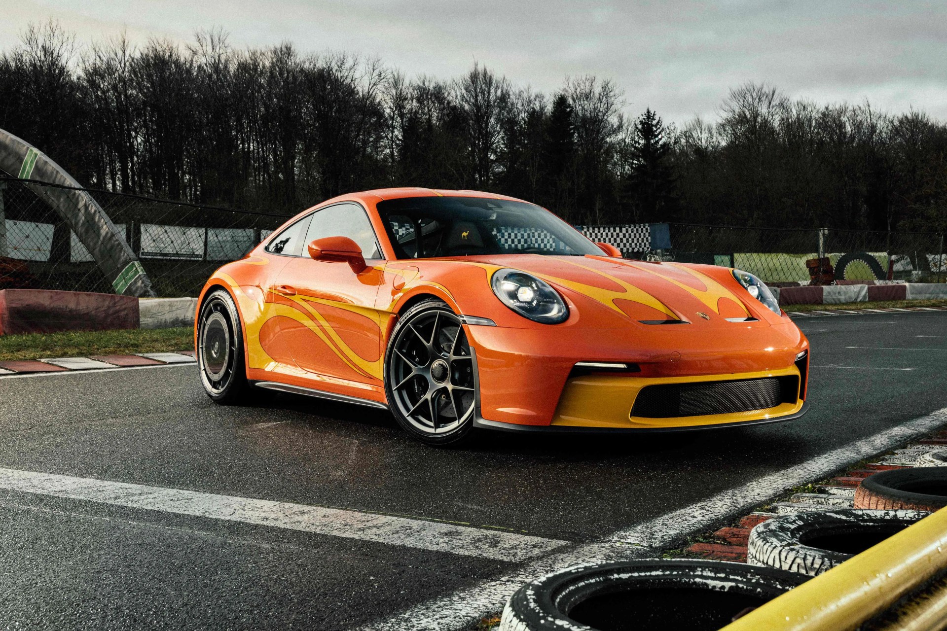 Orange sports car with yellow flame decals parked on a wet race track near tires and barriers.