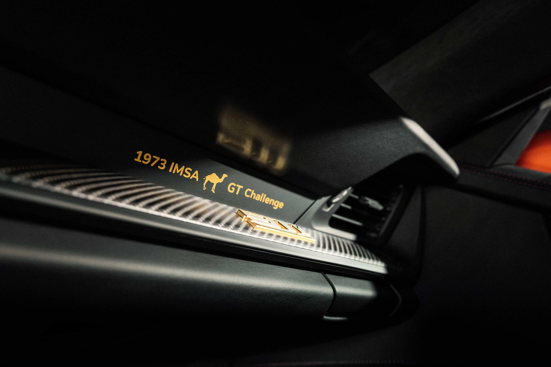 Close-up of a car dashboard with "1973 IMSA GT Challenge" text and a camel logo in gold on a carbon fiber trim.