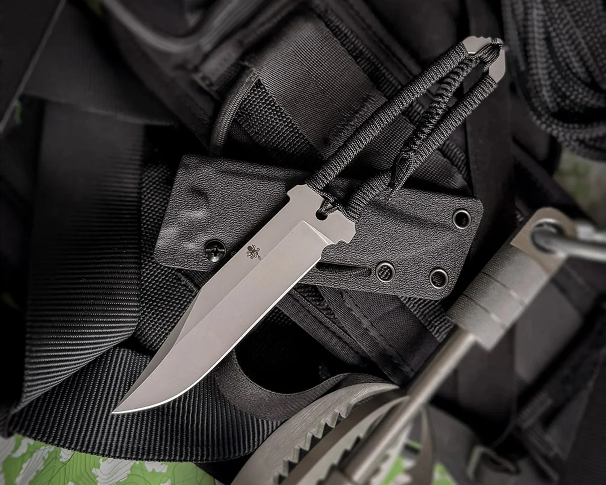 Fixed-blade knife with a silver blade and black paracord-wrapped handle resting on black tactical gear.