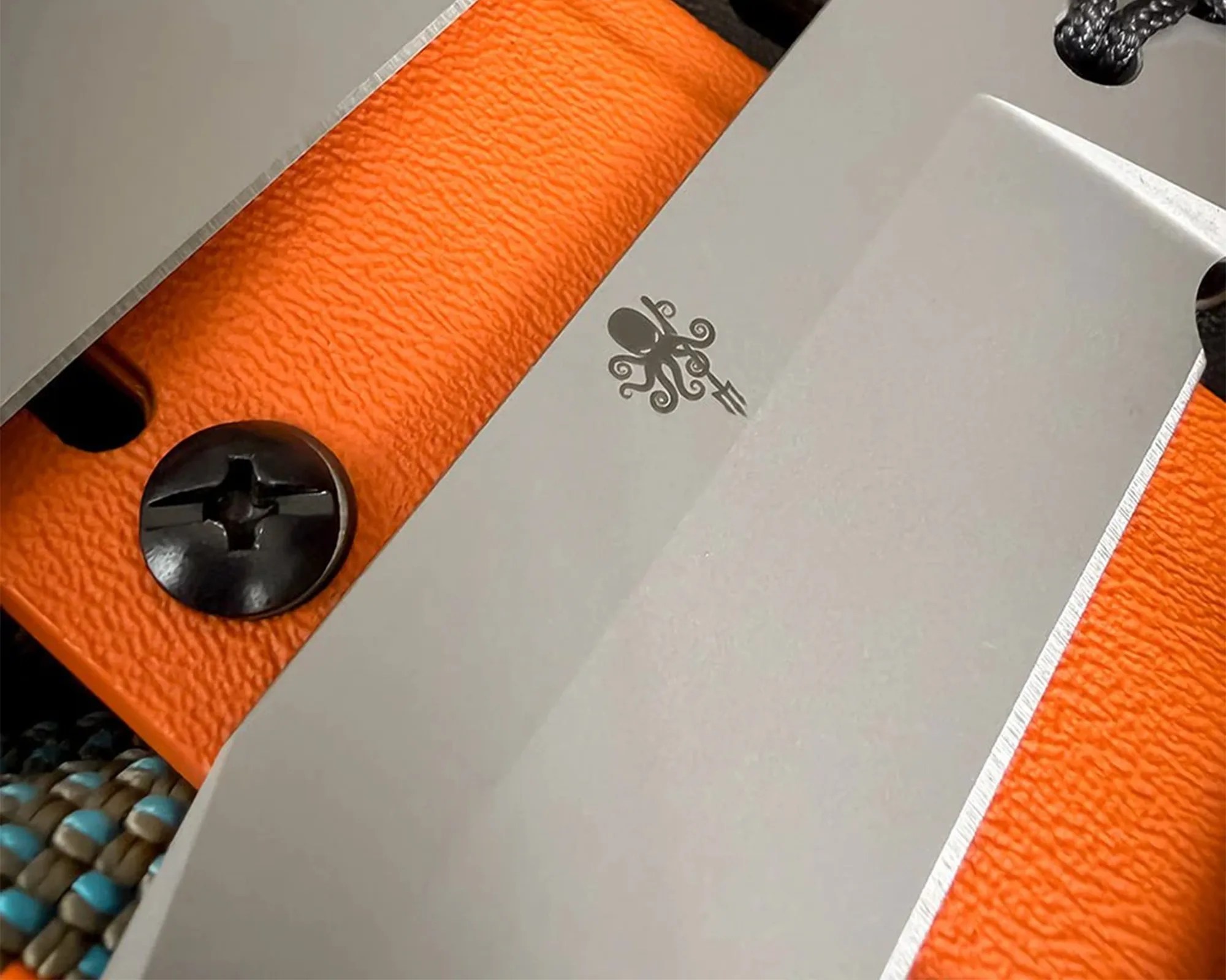 Close-up of a matte silver knife blade with an octopus logo, resting on an orange textured surface with a black screw.