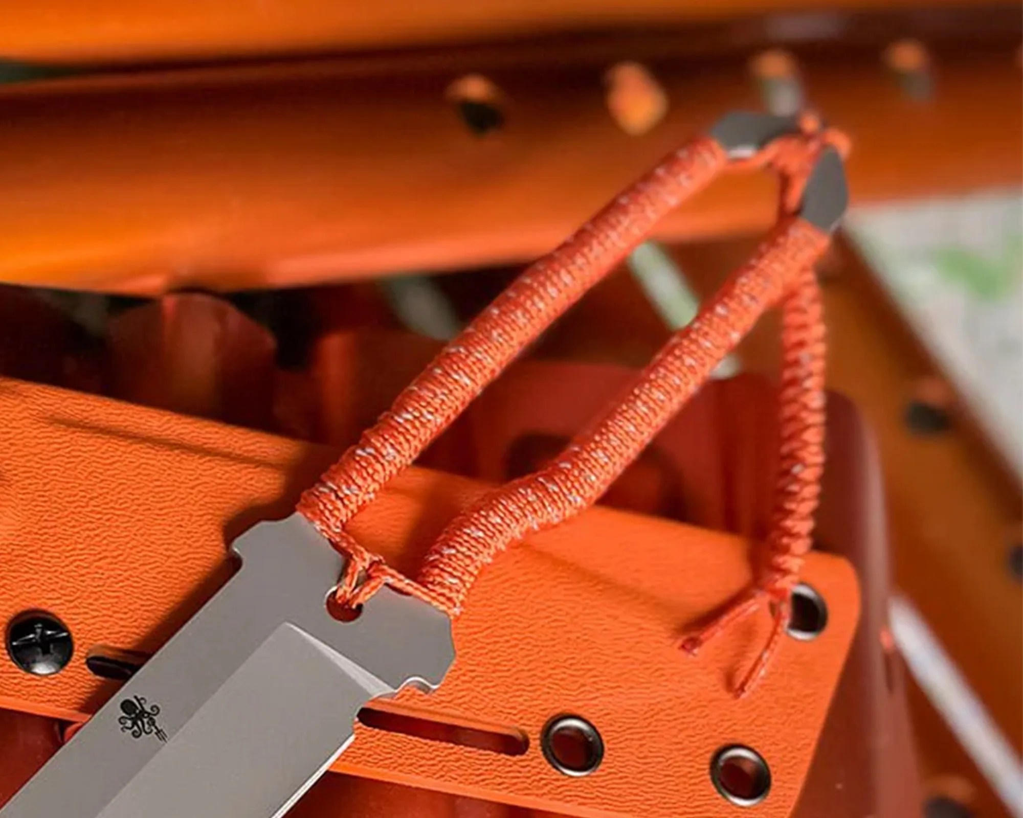 Close-up of a knife with a gray blade and orange paracord-wrapped handle resting on an orange textured sheath.