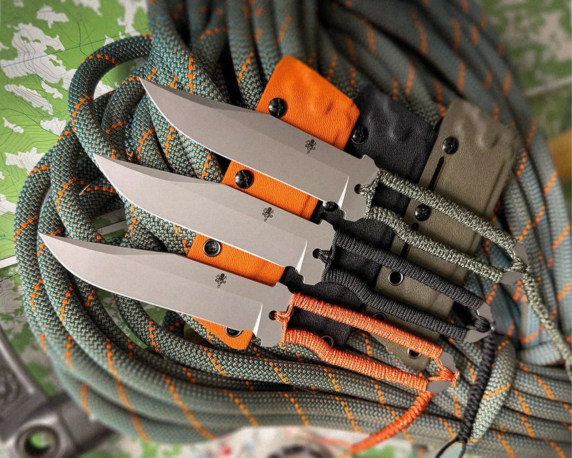Three fixed-blade knives with wrapped handles in green, black, and orange, resting on coiled climbing rope.