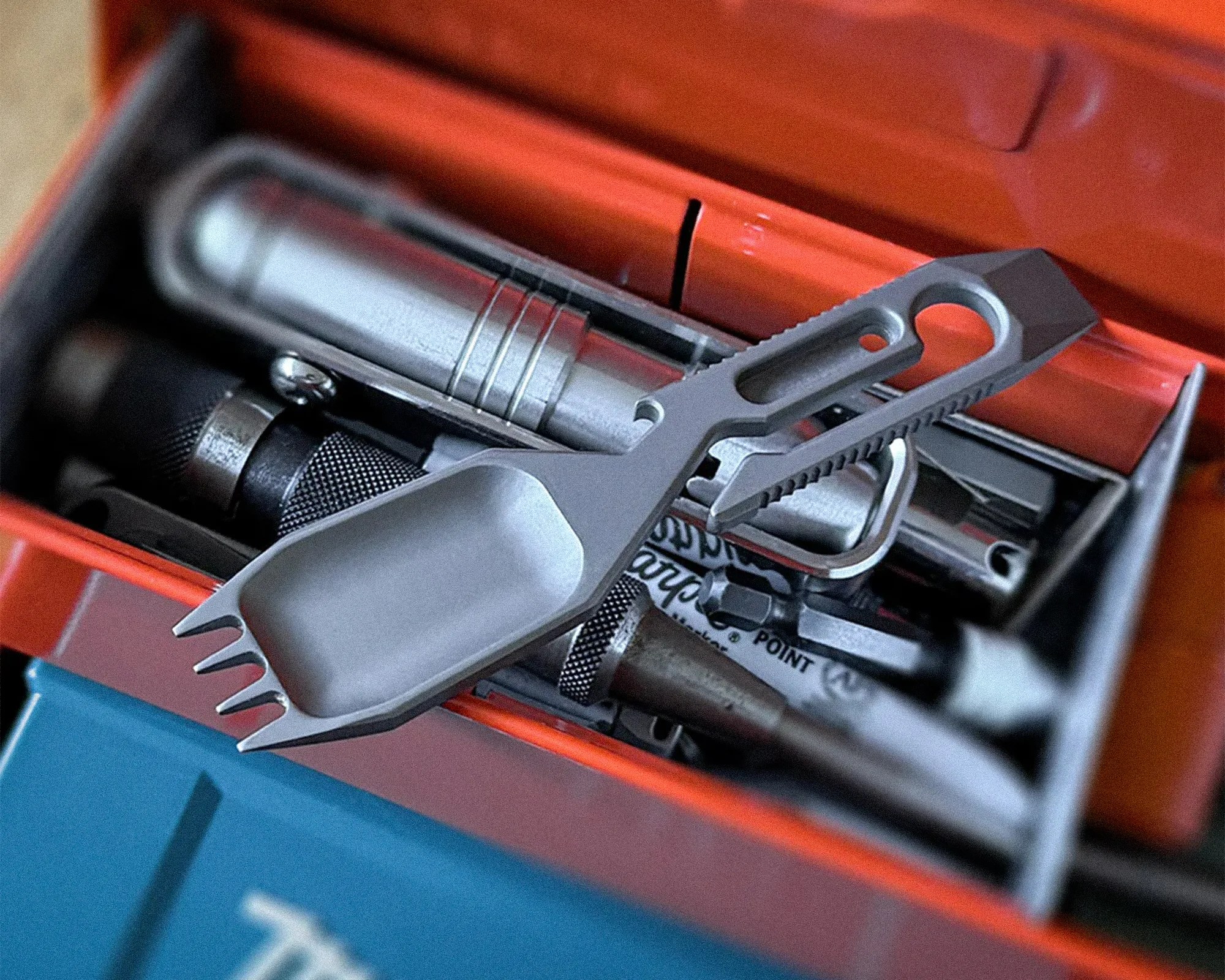 Compact gray metal multi-tool with a scoop, fork, bottle opener, and hex wrench resting on an orange toolbox.