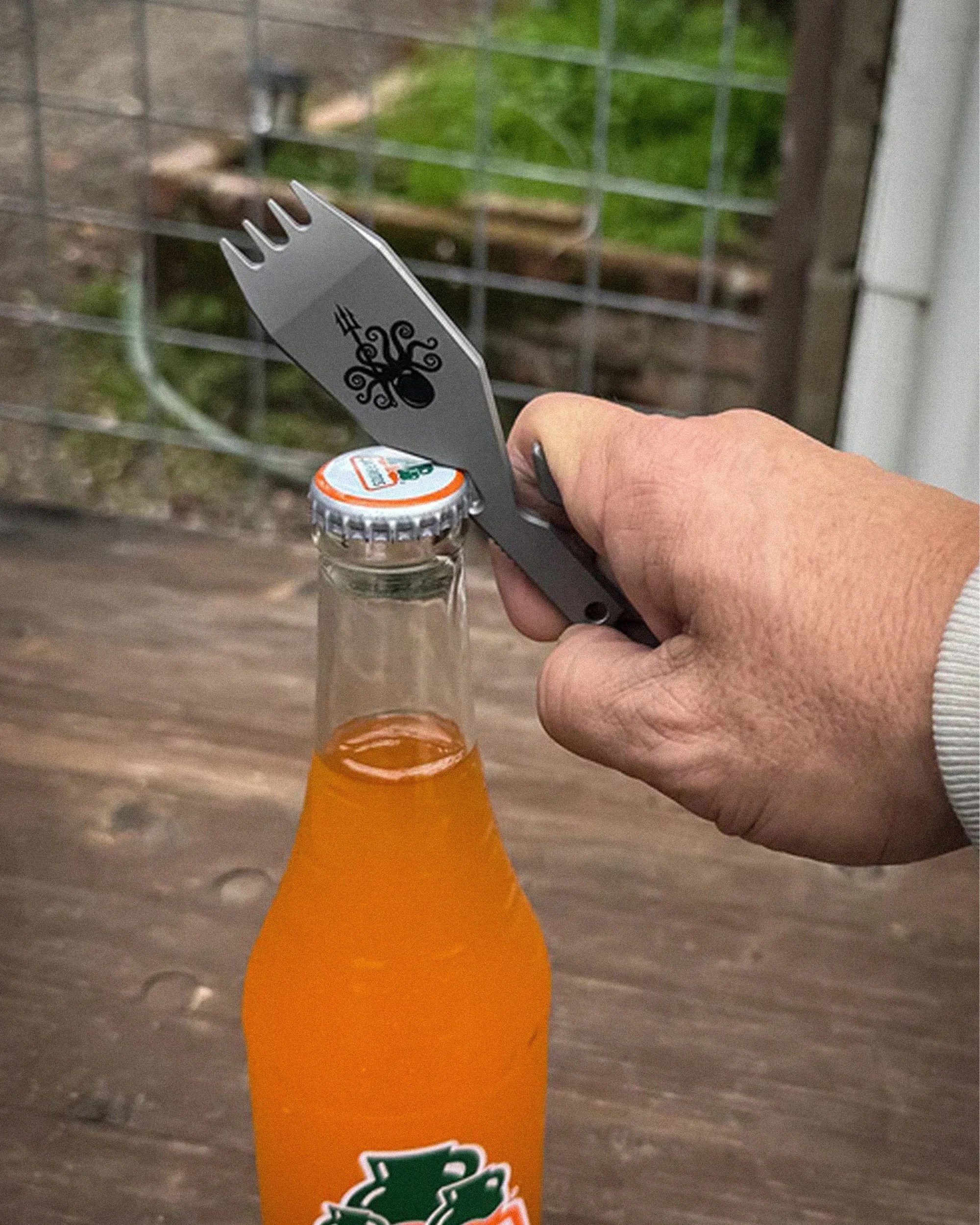 Hand holding a gray metal spork with an octopus logo, using it to open a bottle of orange soda.