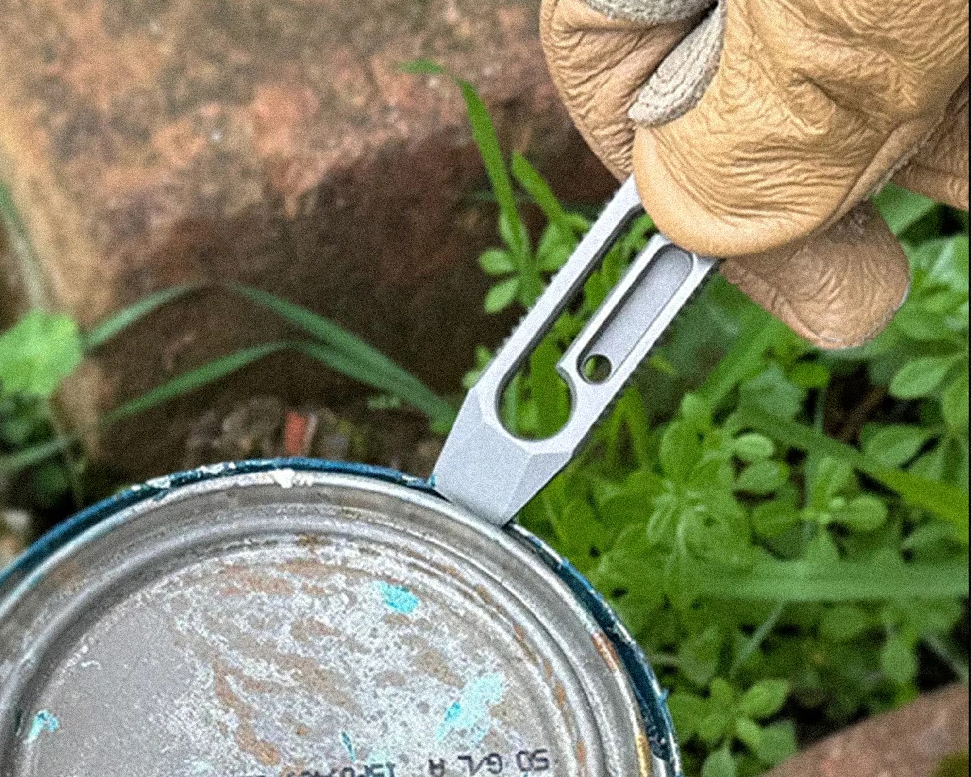 Hand in a tan glove using a small metal tool to pry open a paint can lid outdoors.