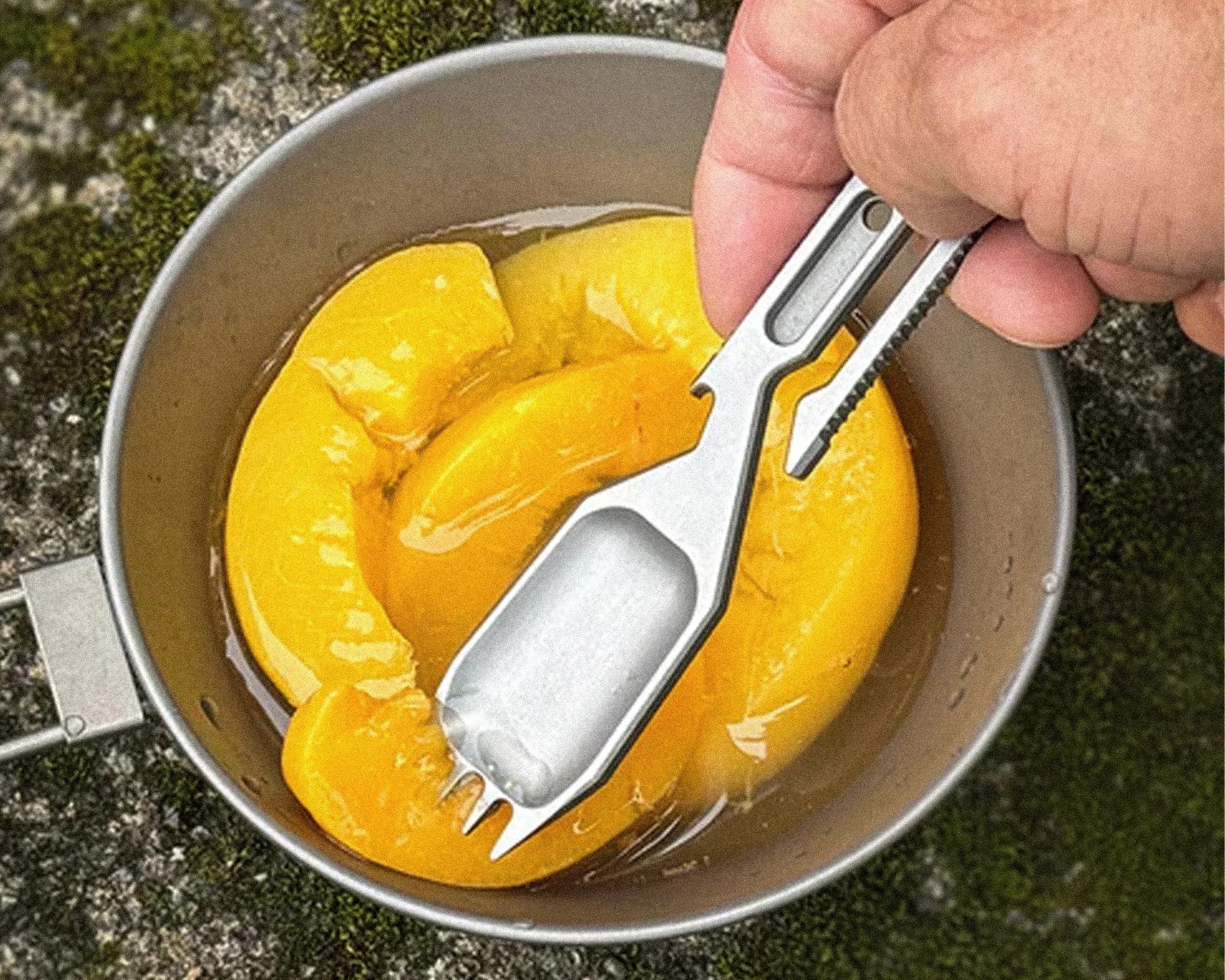Hand holding a metal multi-tool scoop with a serrated edge over a bowl of peeled yellow fruit slices.