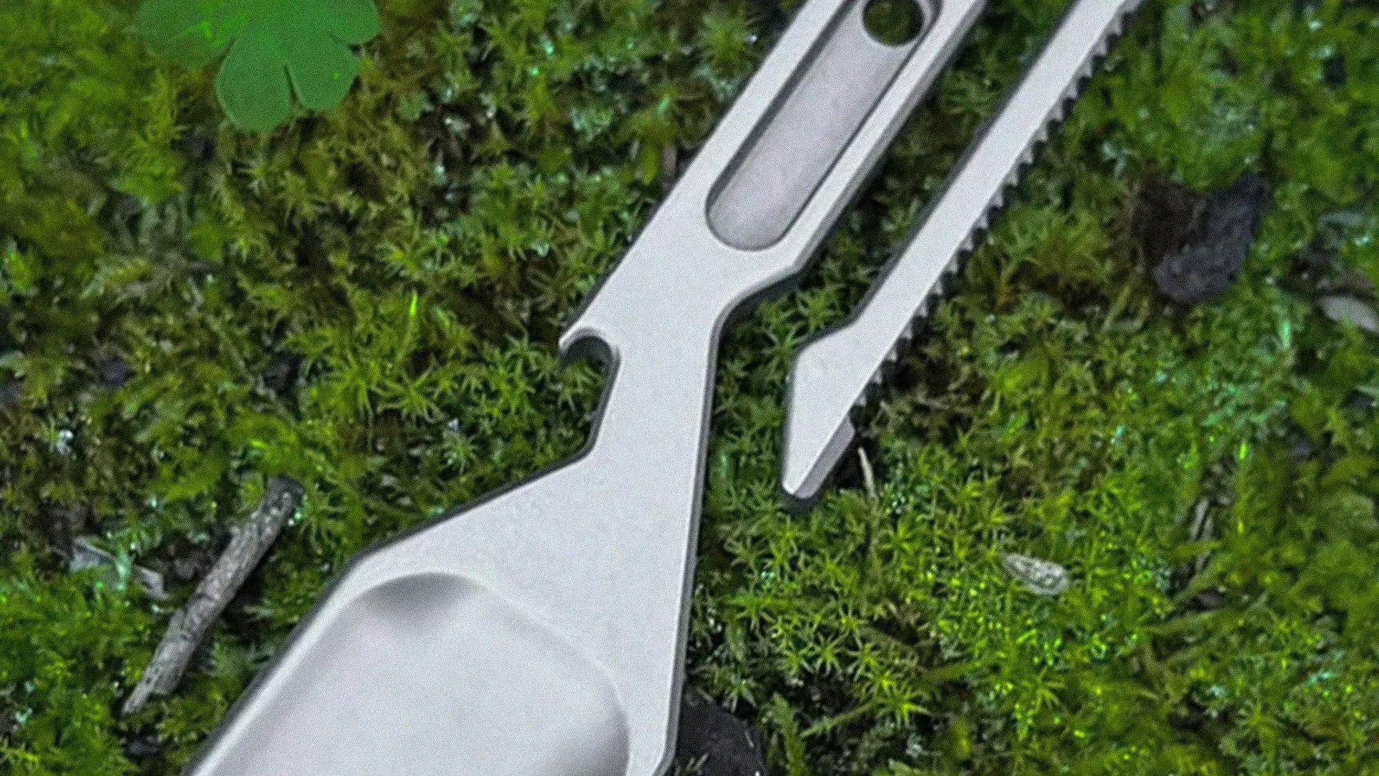 Two silver metal multitools with bottle opener and serrated edge lying on green moss.