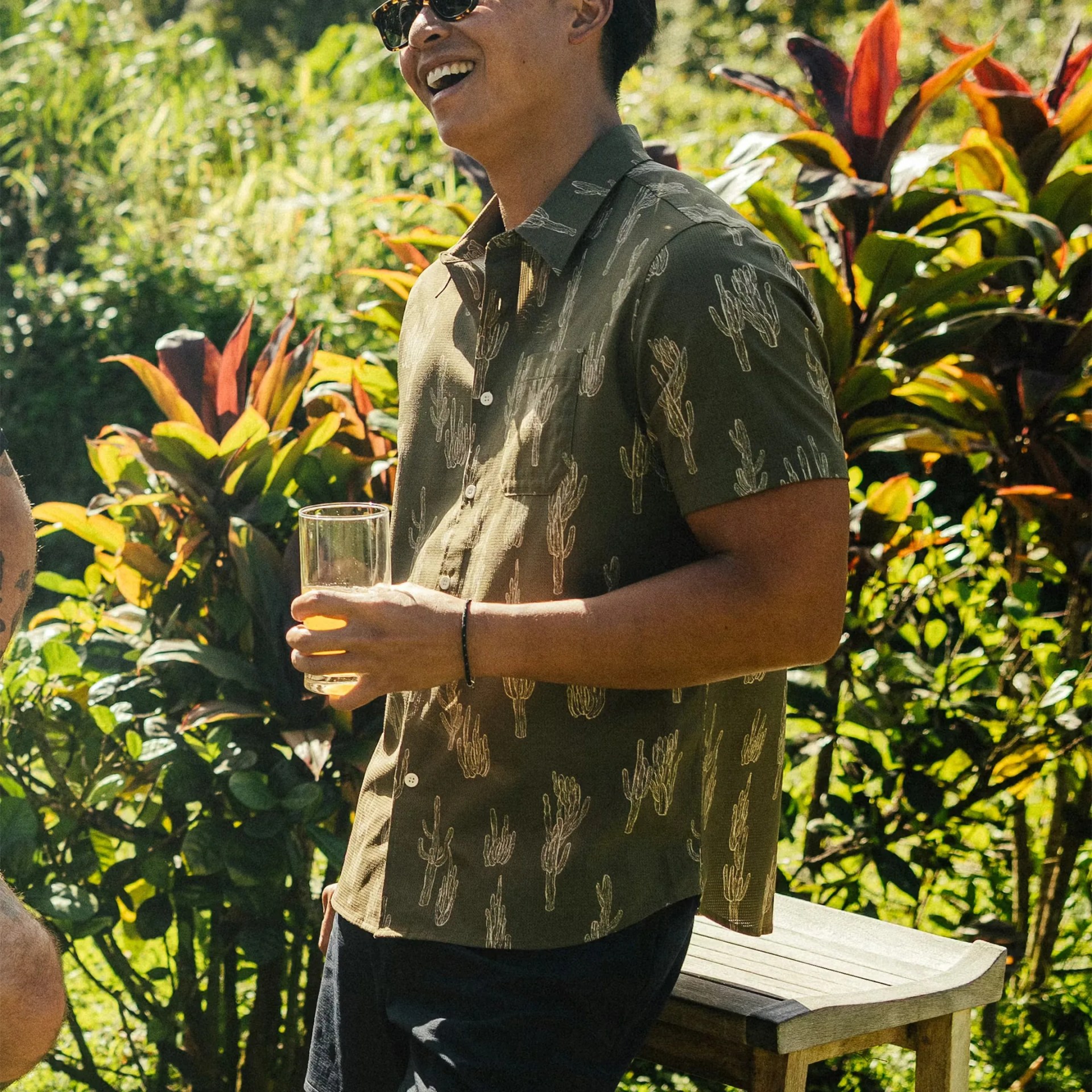 Man wearing an olive green short-sleeve button-up shirt with white cactus prints holding a glass outdoors.
