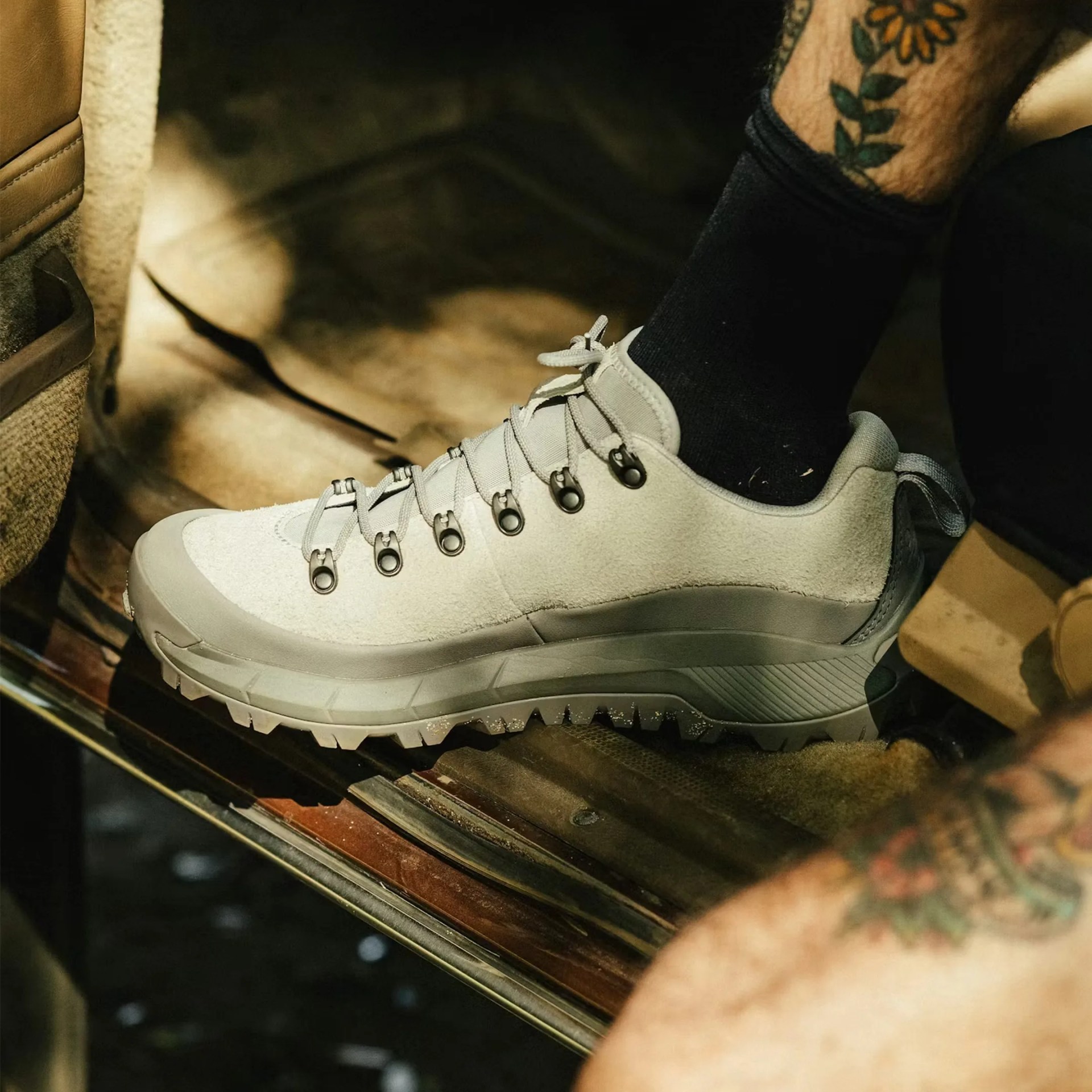Light gray suede hiking shoe with black metal eyelets and rugged sole worn by a person with tattoos.