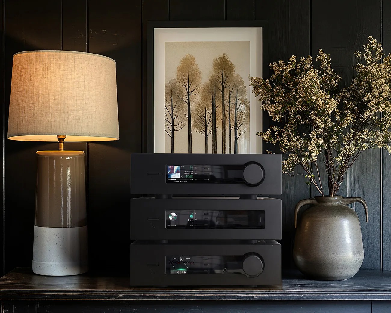 Stack of three black audio components with digital displays on a dark wooden surface, next to a beige and white ceramic lamp and a large gray vase with flowering branches.
