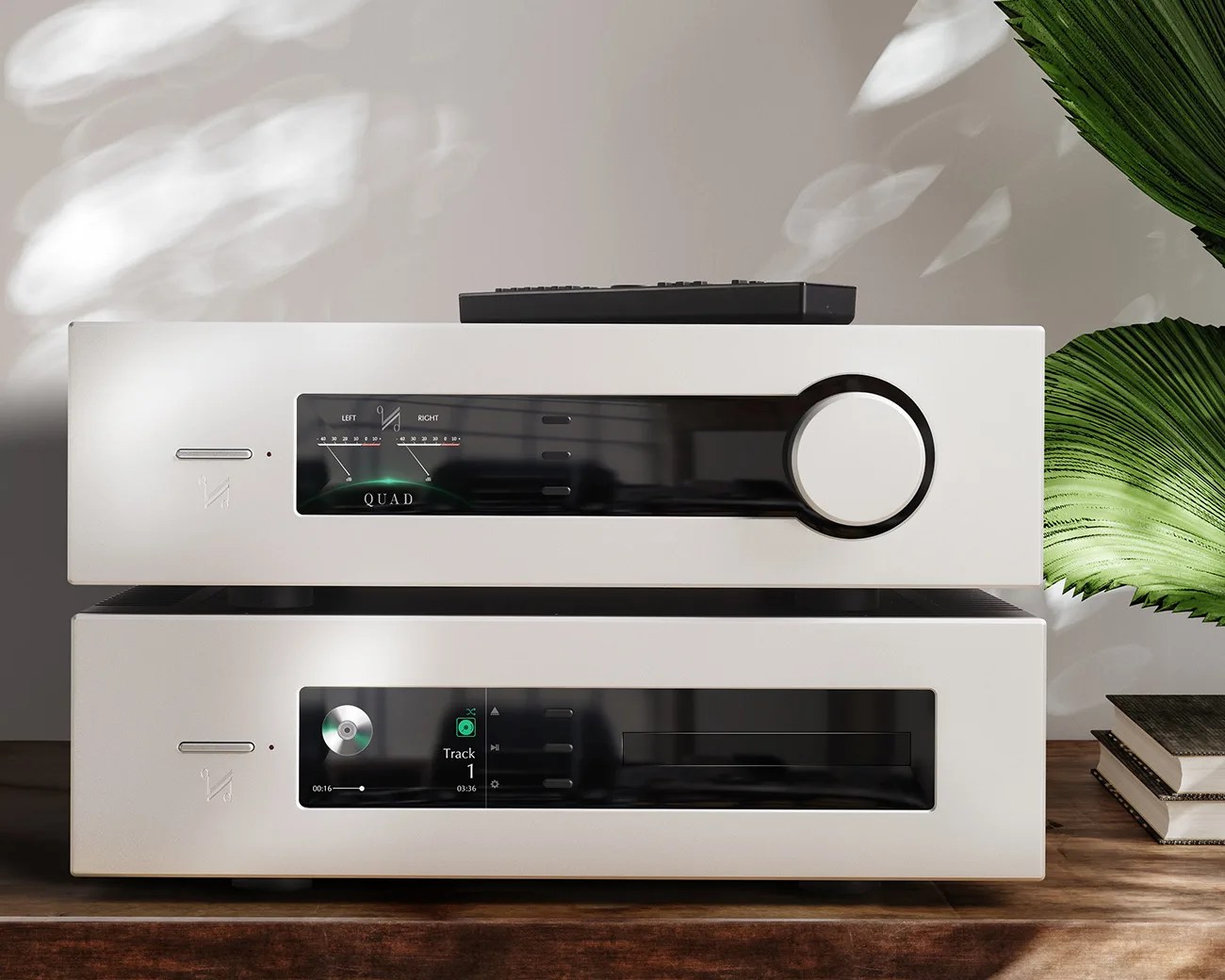 Two stacked white audio components with black display panels, a large knob on the top unit, and a remote control on top, placed on a wooden surface next to green leaves and books.
