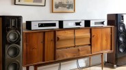 Pair of large black floor-standing speakers flanking a mid-century wooden sideboard with three silver audio components on top.