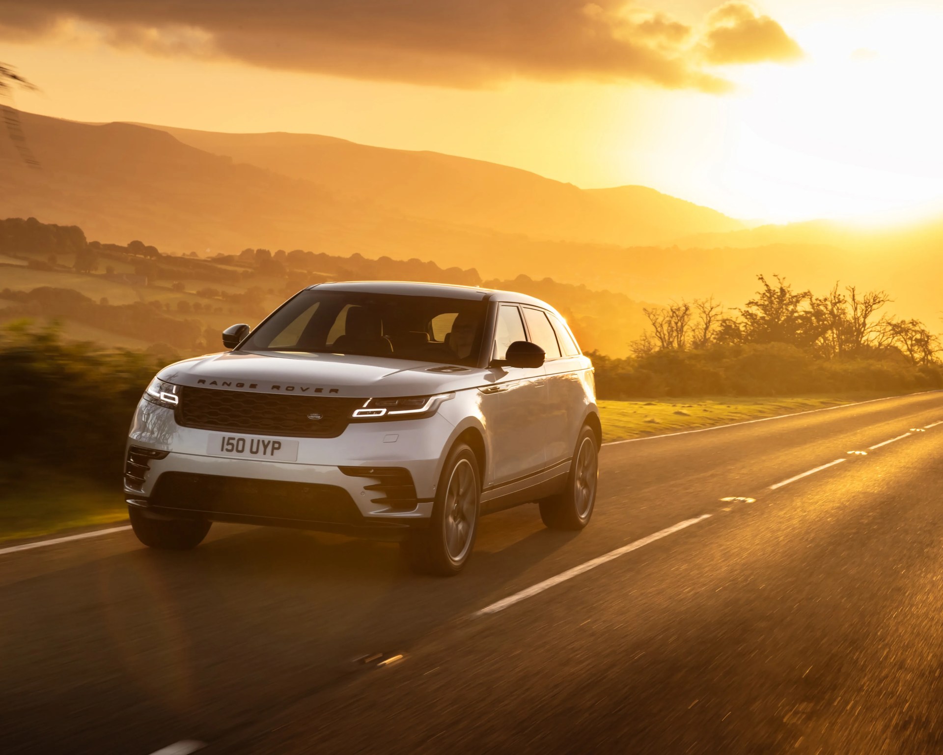 Silver Range Rover SUV driving on a rural road at sunset with hills in the background.