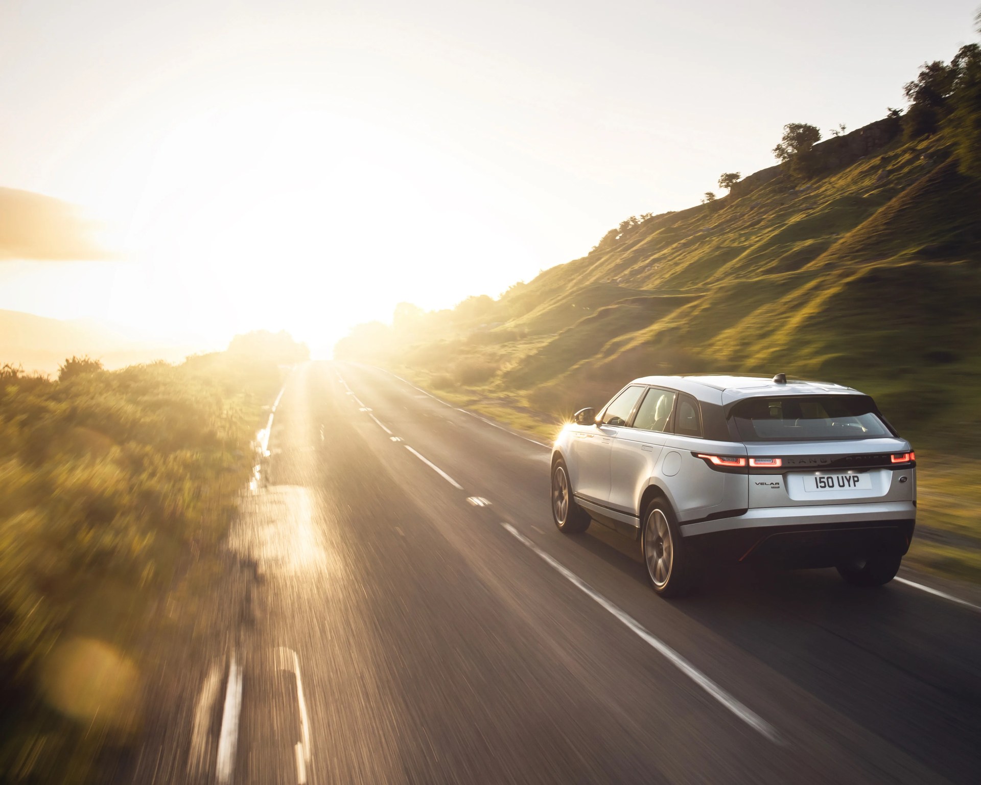 Silver Range Rover Velar driving on a sunlit rural road with green hills on the right.