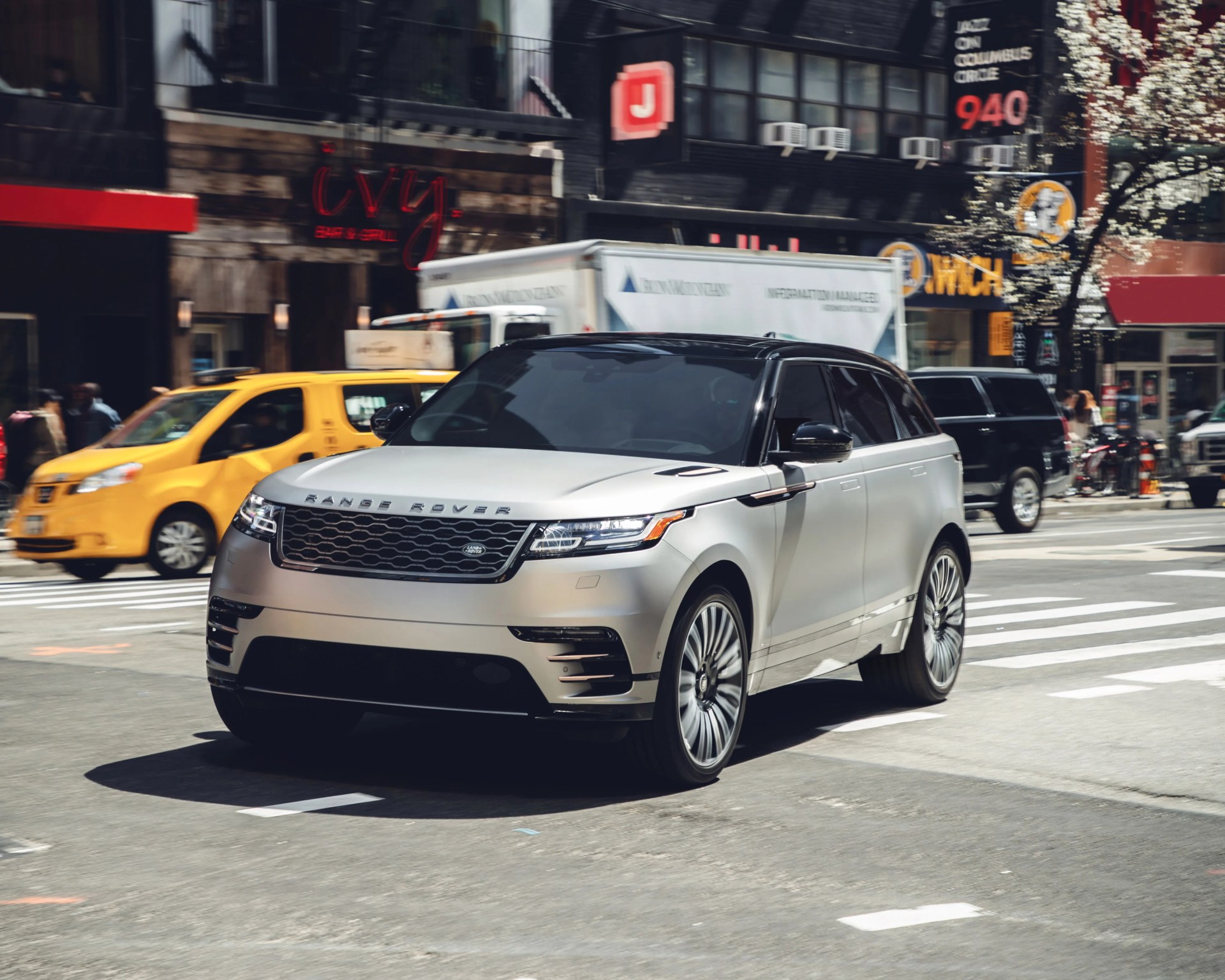 Silver Range Rover SUV driving on a city street with a yellow taxi and delivery truck in the background.