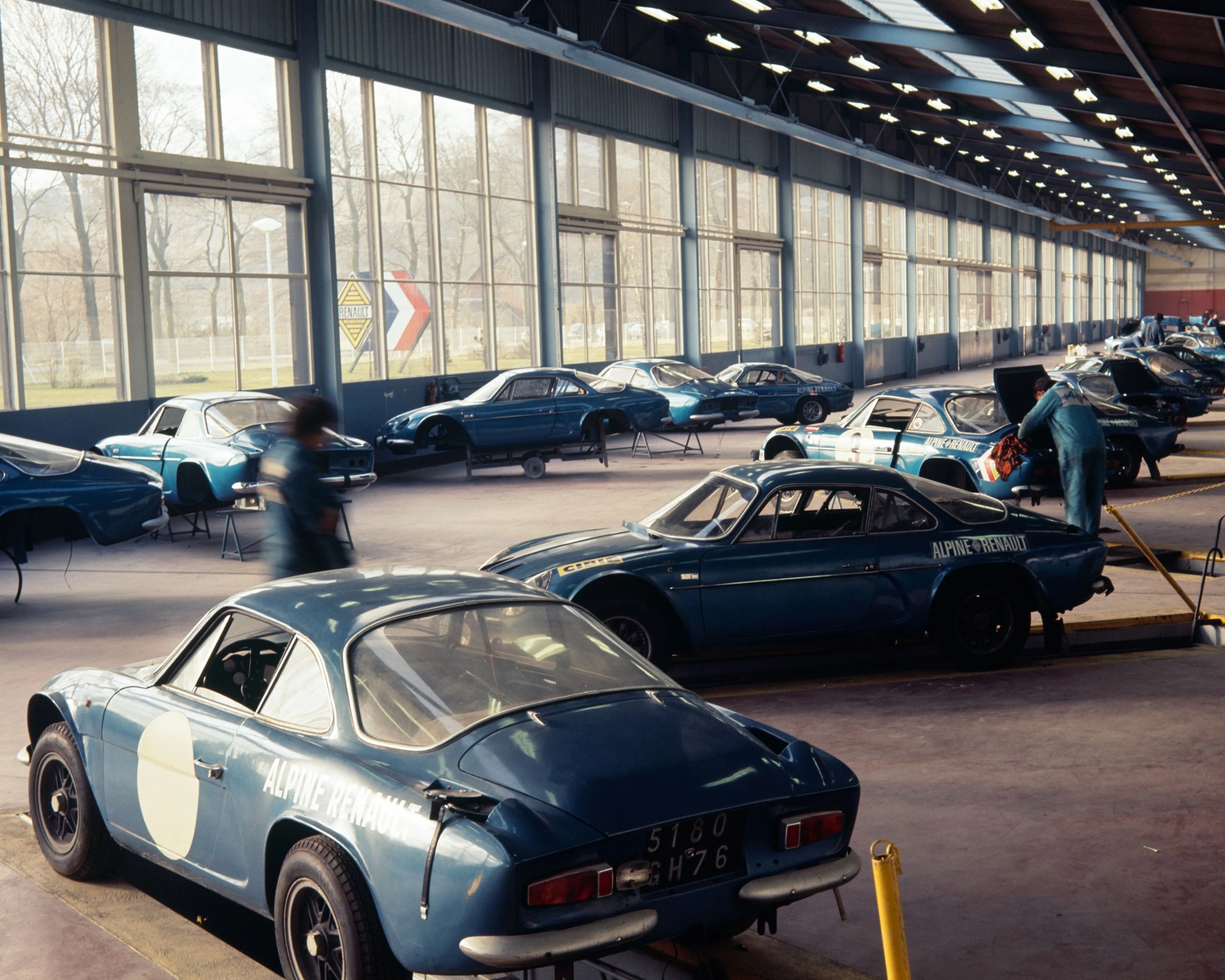 Blue Alpine Renault race cars in a large workshop with mechanics working on them.