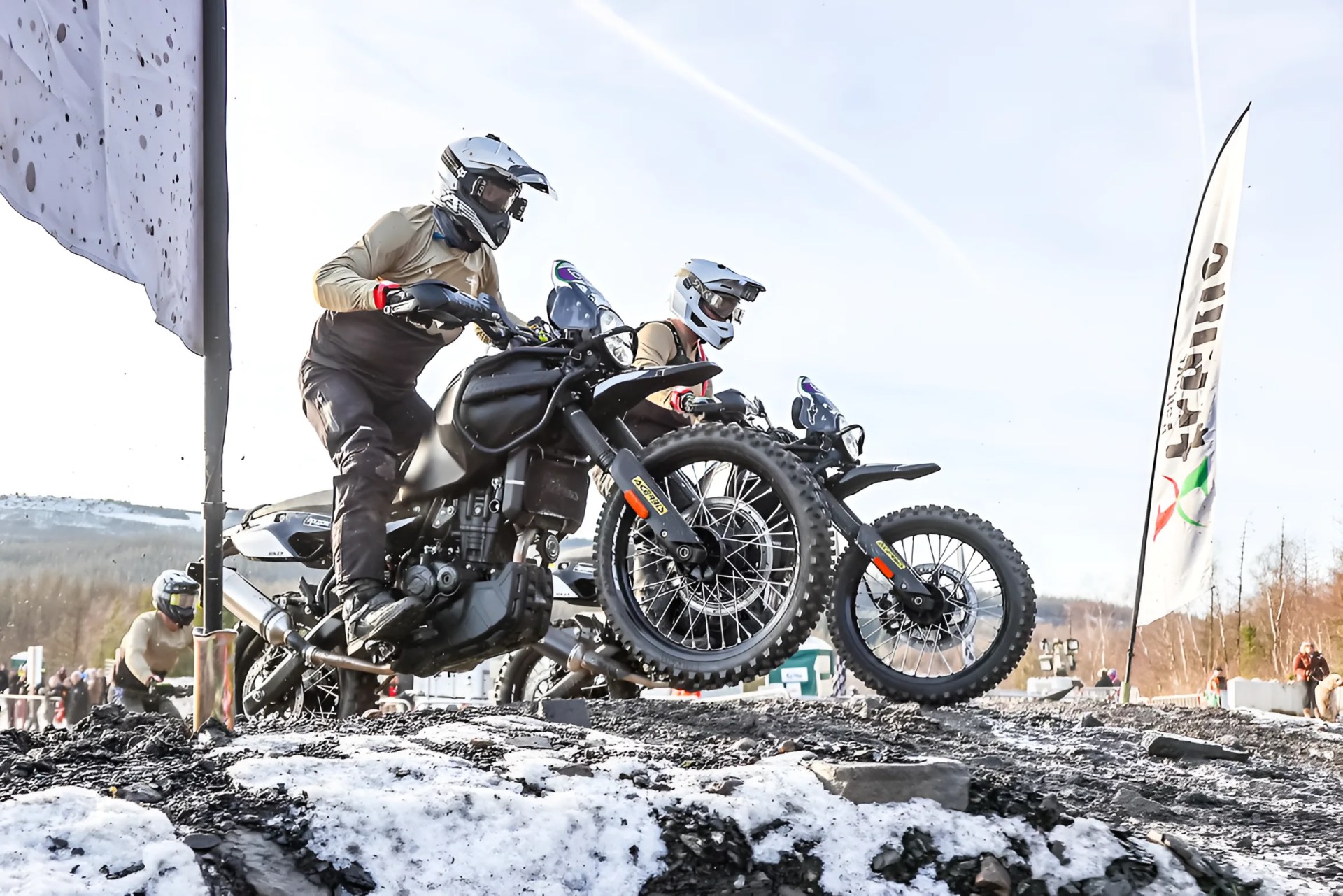 Two riders in helmets on black dirt bikes jumping over a snowy, muddy hill with flags in the background.