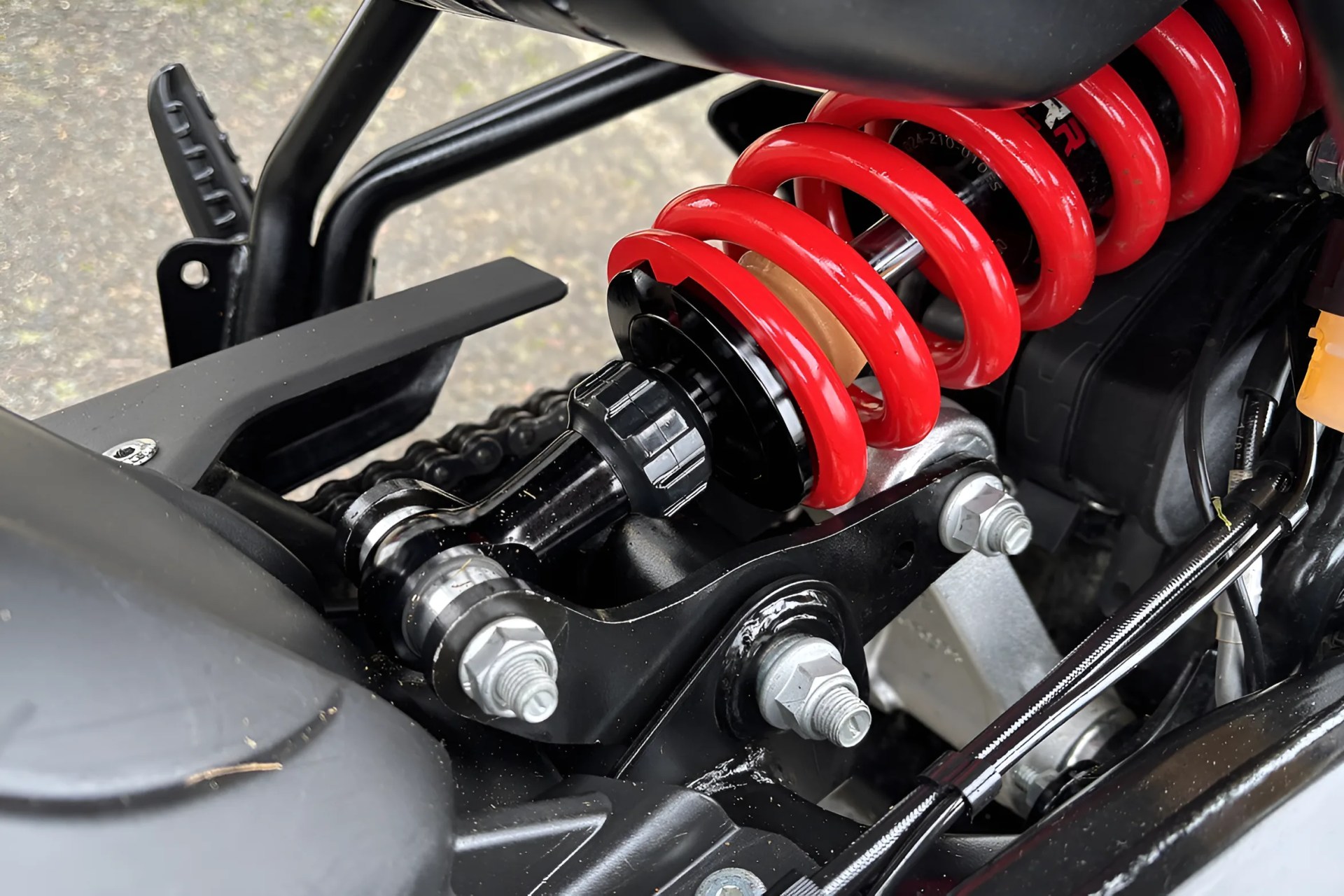 Close-up of a red coil spring shock absorber mounted on a black motorcycle frame near the chain and footrest.