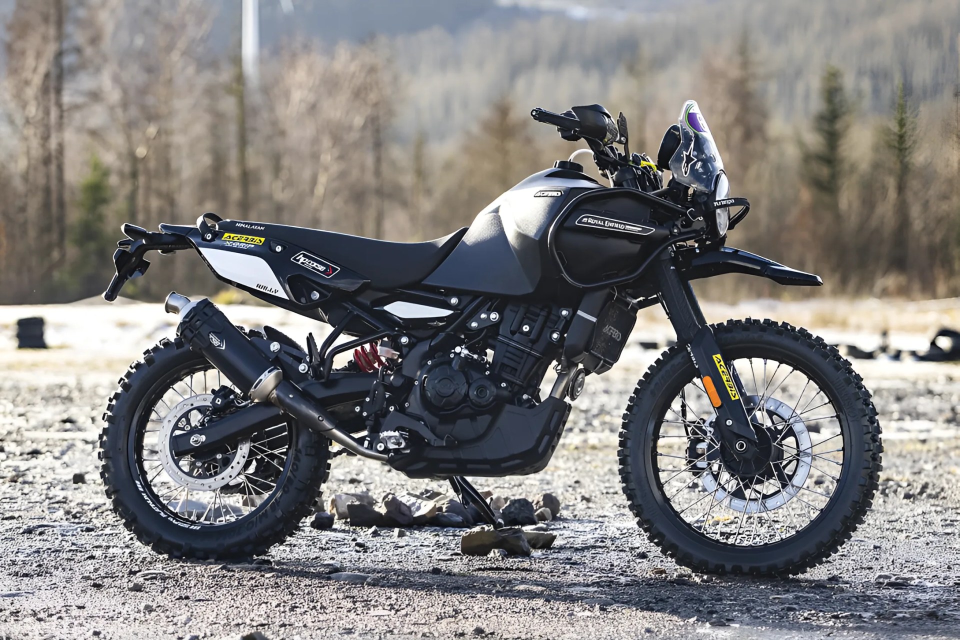 Black off-road motorcycle with knobby tires and a small windshield parked on gravel with a forest background.