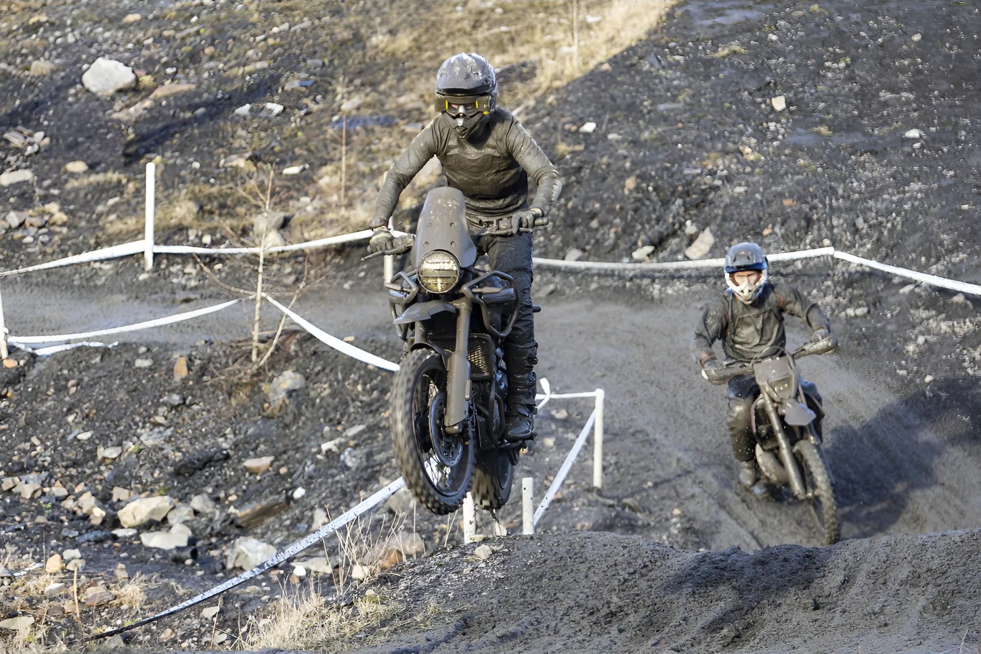 Two motorcyclists wearing helmets and dark gear riding dirt bikes on a rocky off-road trail, one airborne over a small jump.