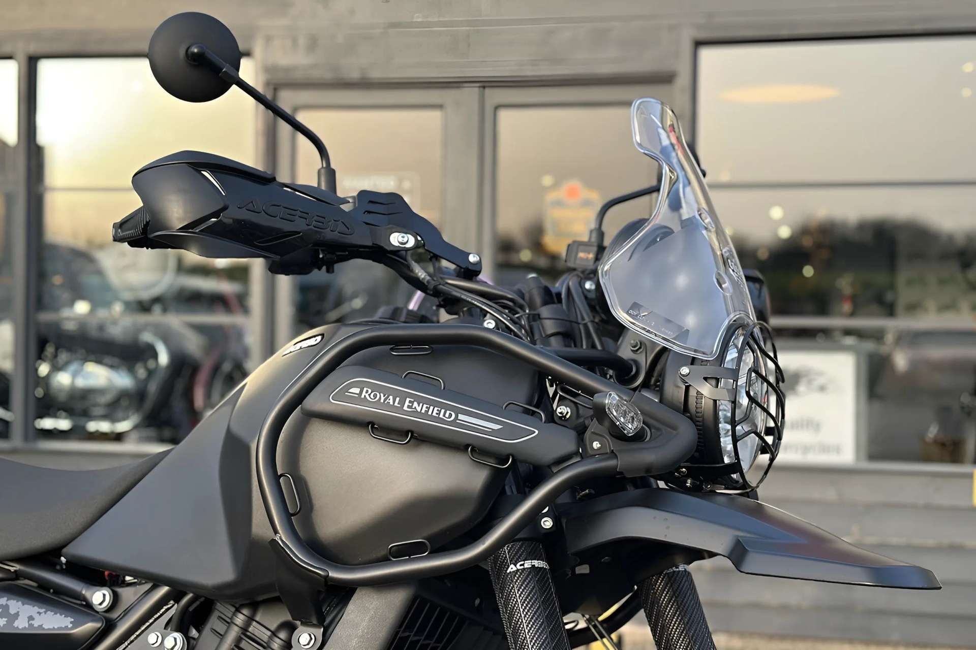 Matte black Royal Enfield motorcycle with handguard, crash bars, and clear windscreen in front of a building.