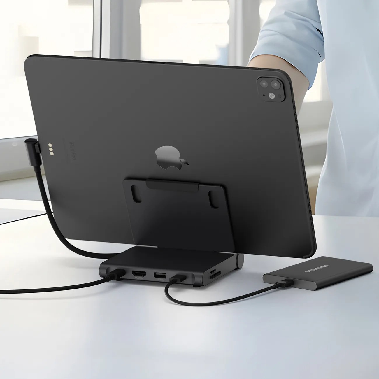 Black iPad Pro mounted on a black multi-port hub with cables connected, next to a Samsung external SSD on a white table.