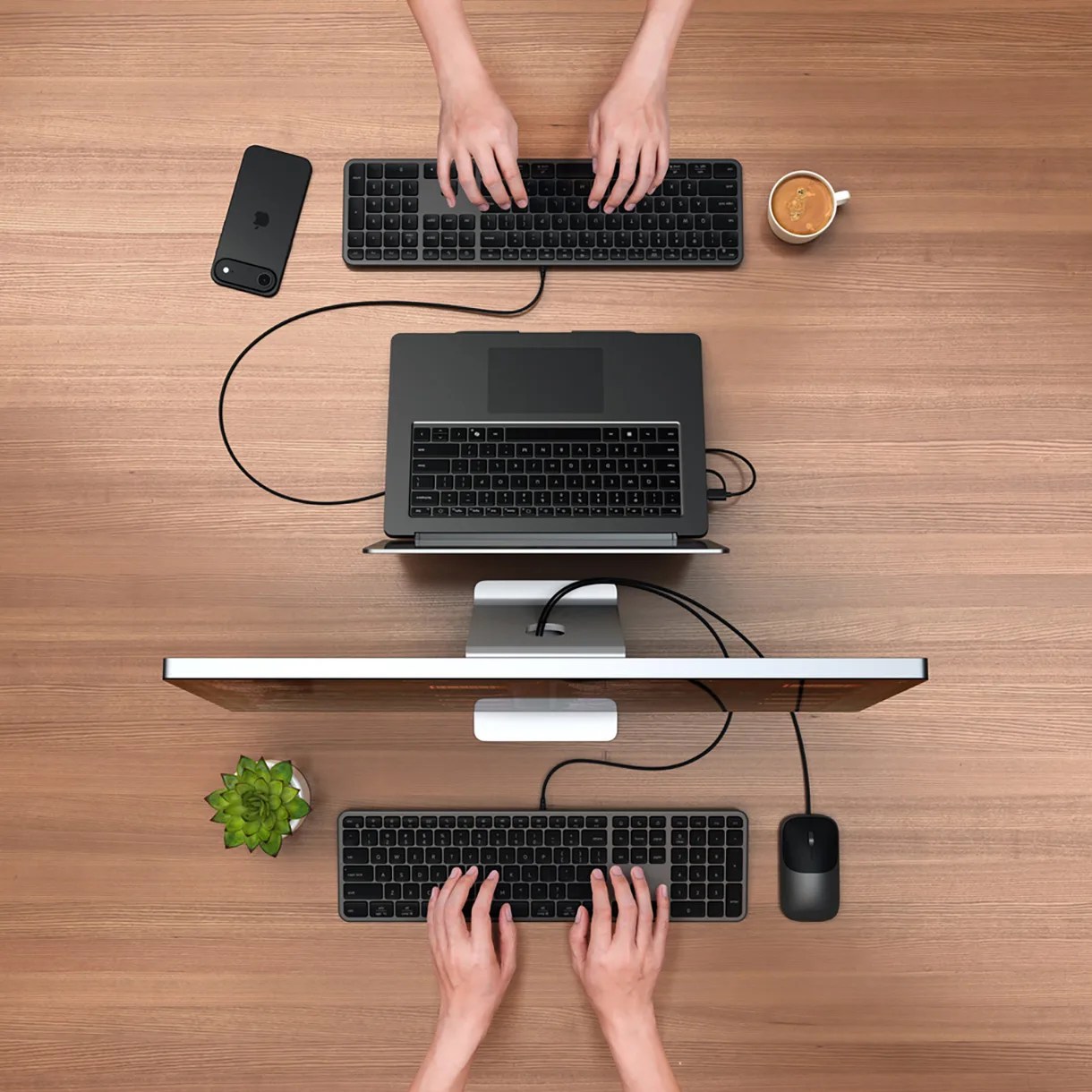 Top-down view of a wooden desk with two sets of hands typing on black keyboards, a black mouse, a black laptop connected to a monitor, a black smartphone, a cup of coffee, and a small green succulent plant.