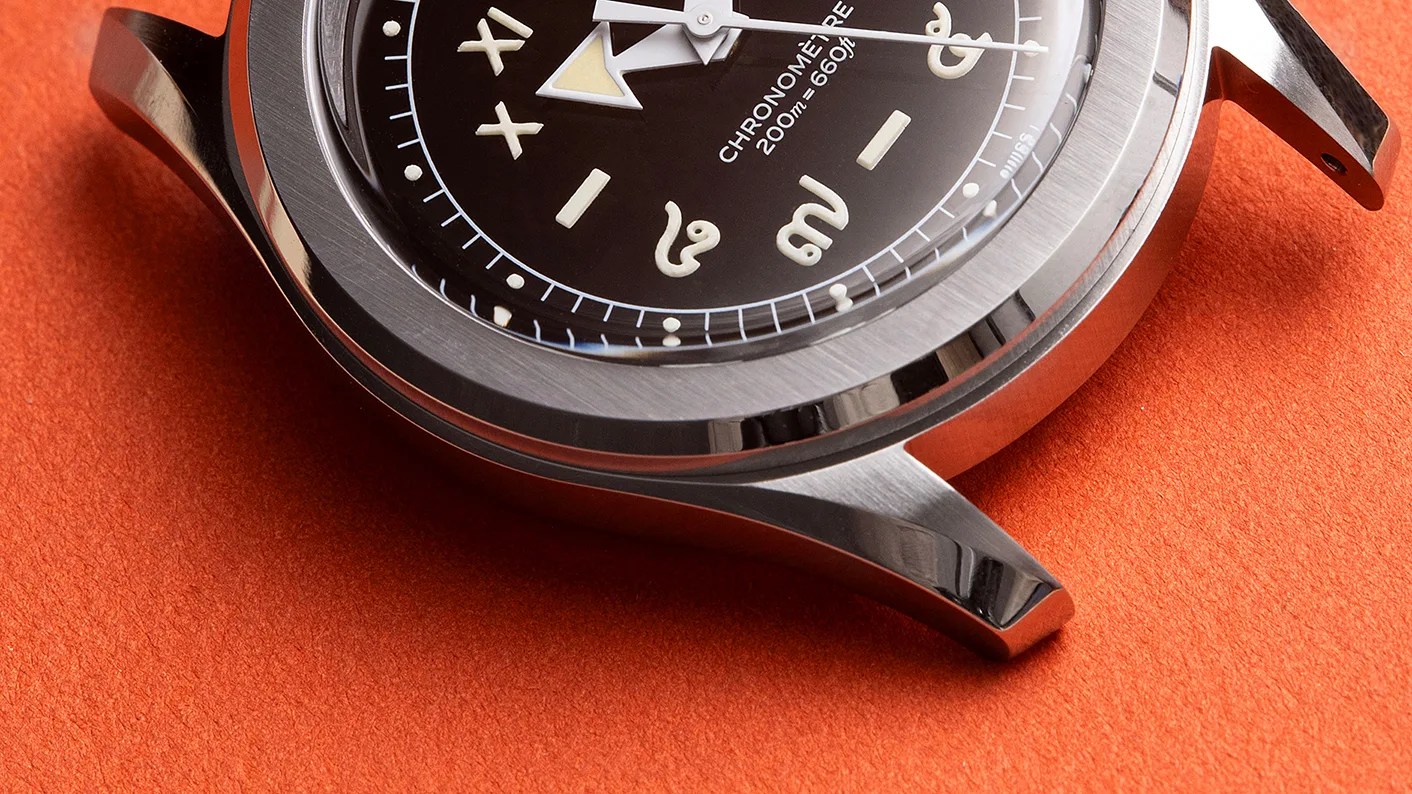 Close-up of a stainless steel watch case with a black dial featuring luminous markers and text "CHRONOMÈTRE 200m=660ft" on an orange surface.