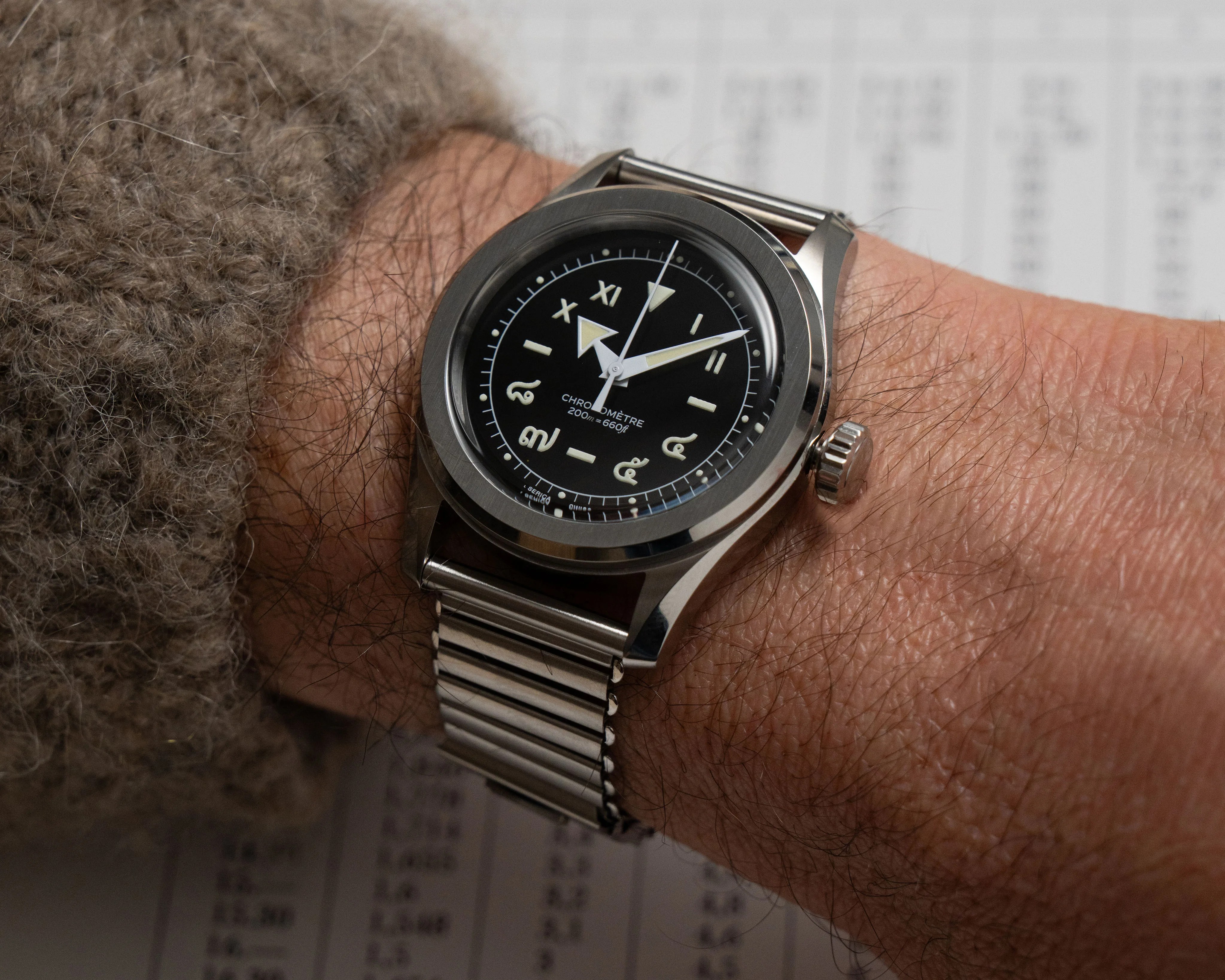 Stainless steel wristwatch with black dial and luminous hands on a wrist wearing a brown knit sleeve.