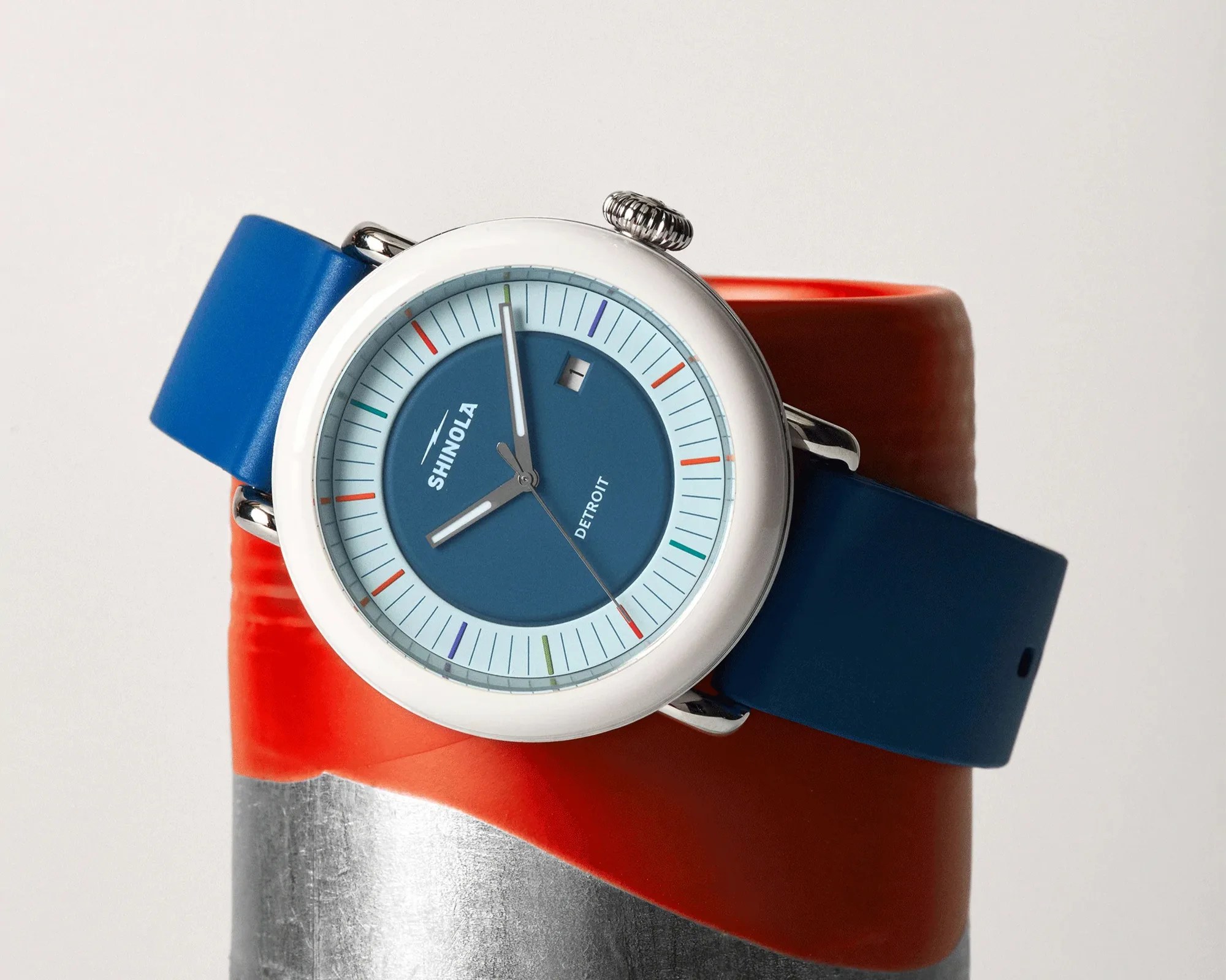 Round Shinola watch with blue dial, multicolored minute markers, date window, and blue silicone strap on red and silver surface.