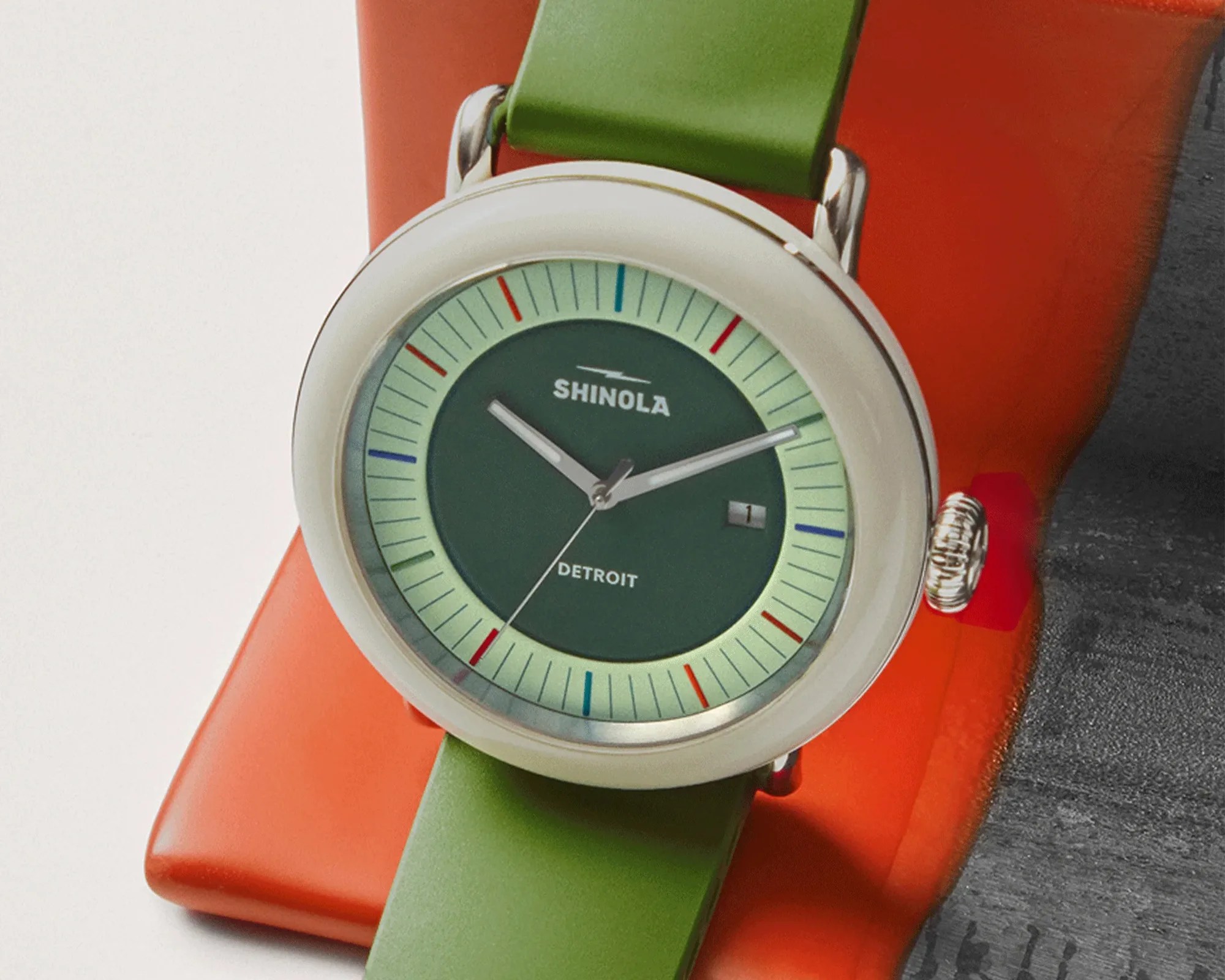 Round Shinola wristwatch with green dial, light green outer ring, silver case, and green strap on an orange stand.