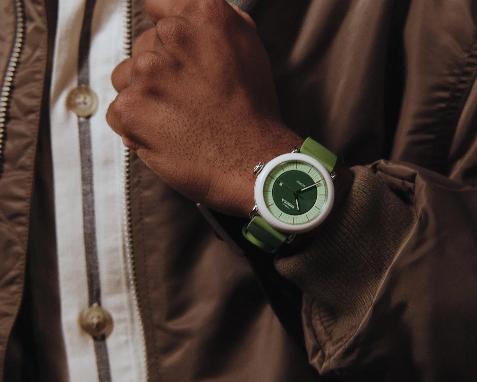 Wristwatch with a green dial and strap, white bezel, worn on a wrist over a brown jacket sleeve.