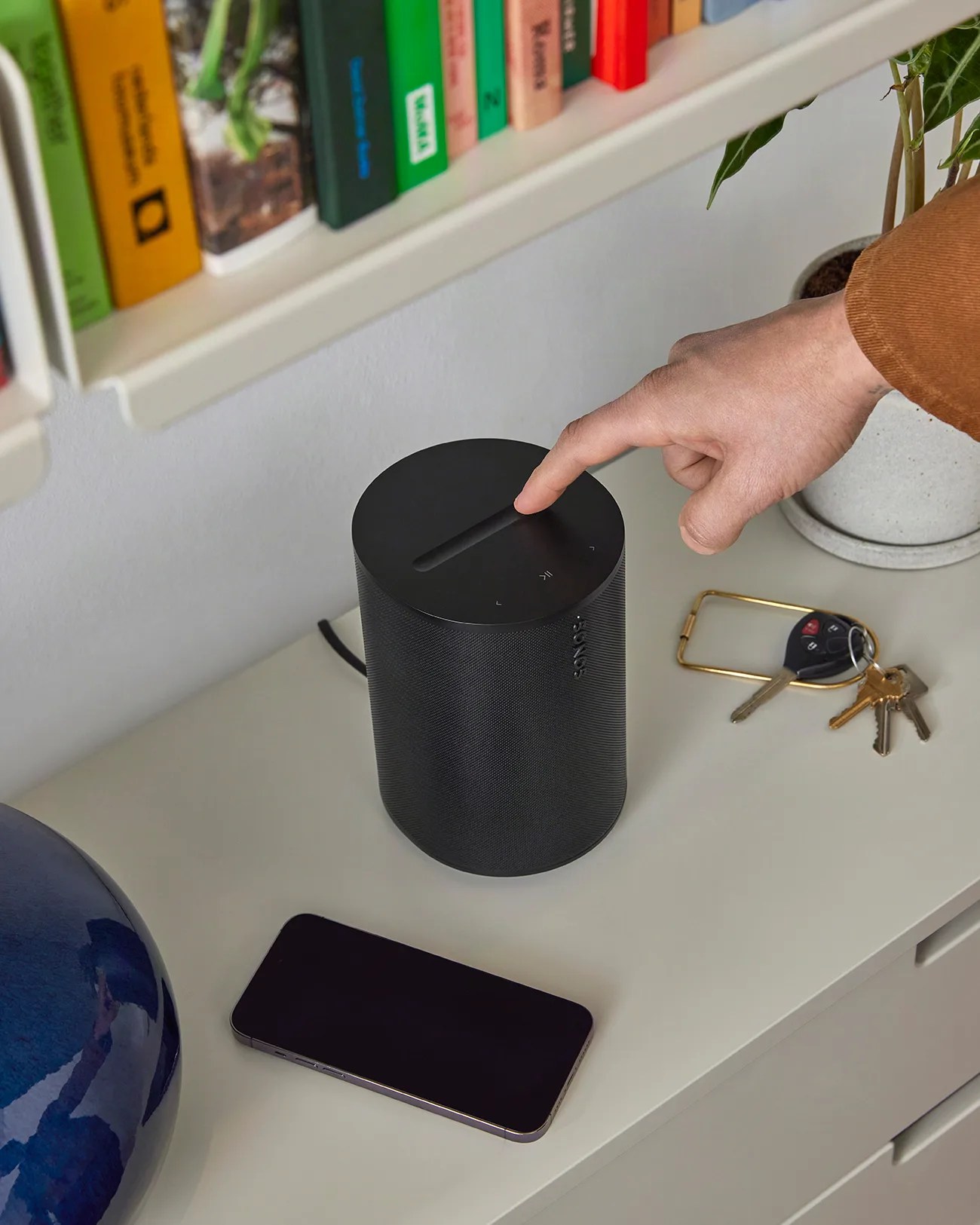 Black cylindrical Sonos speaker on a white surface with a hand adjusting the top controls.