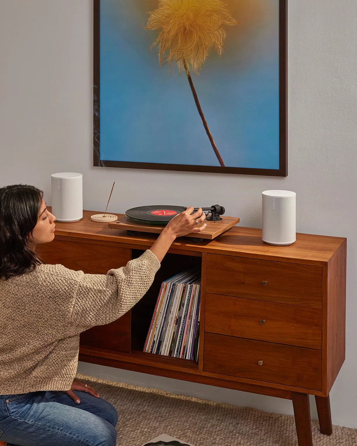 Person adjusting a wooden turntable on a mid-century modern wooden cabinet with white Sonos speakers and vinyl records.