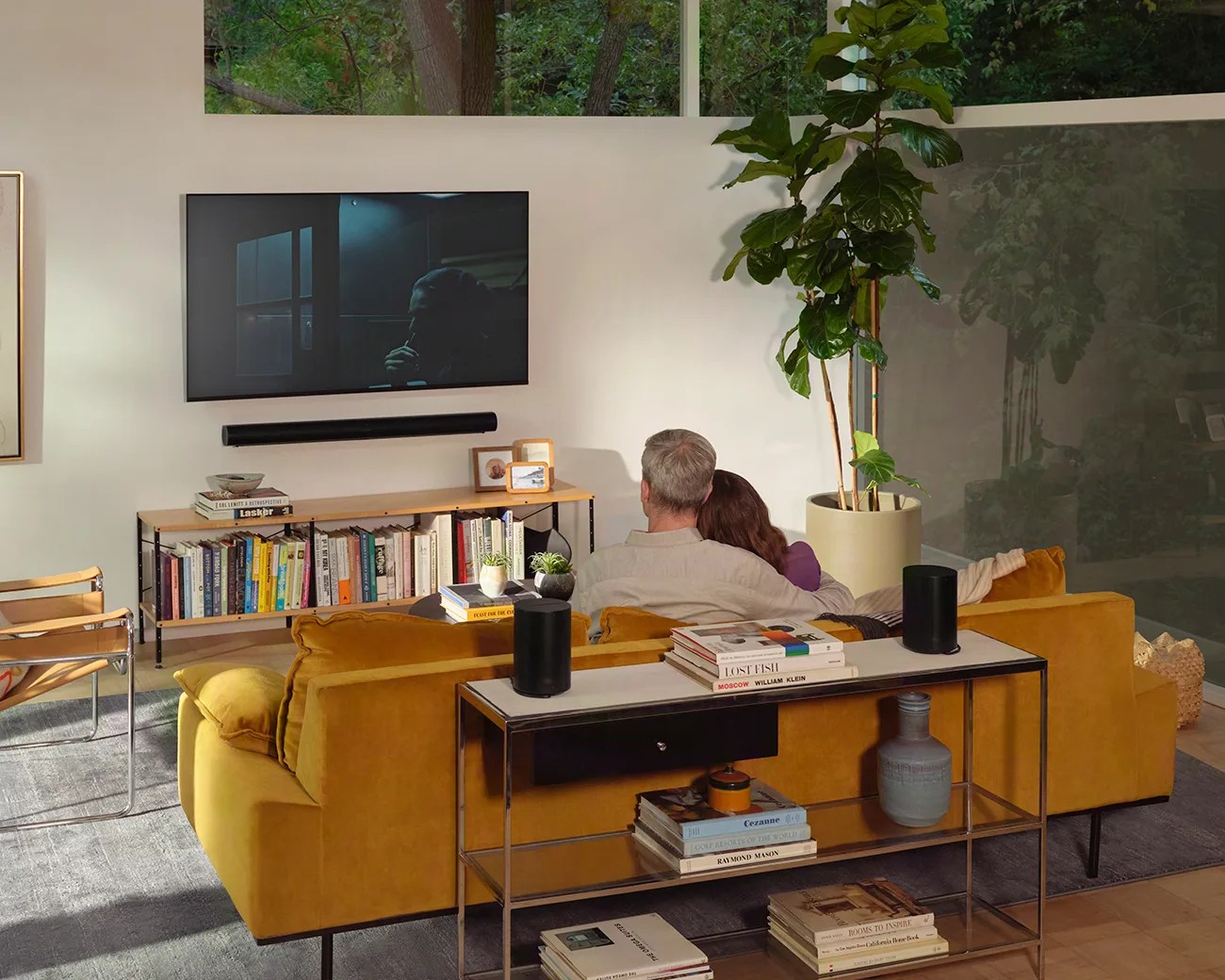 Couple sitting on a mustard yellow sofa facing a wall-mounted TV in a modern living room with bookshelves and a large potted plant.