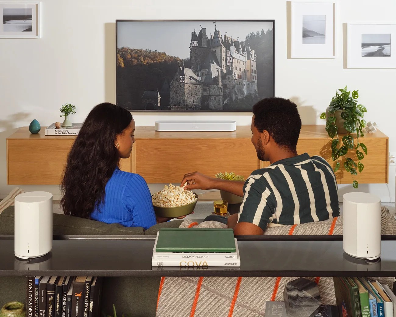 Two people sitting on a couch with a bowl of popcorn, facing a TV mounted above a wooden cabinet with a white soundbar.