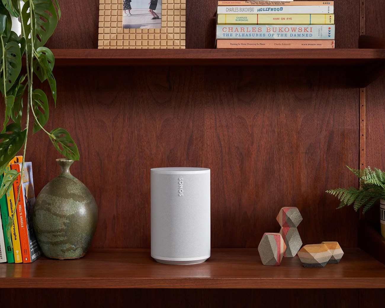 White Sonos speaker on a wooden shelf surrounded by books, a green vase, and geometric decorative objects.