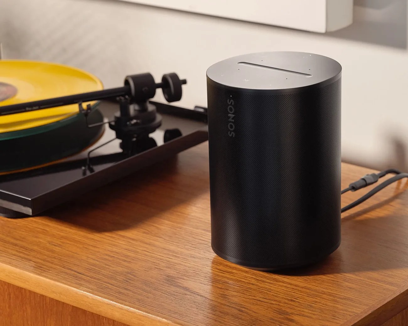 Black cylindrical Sonos speaker with touch controls on top, placed on a wooden surface near a yellow vinyl record player.